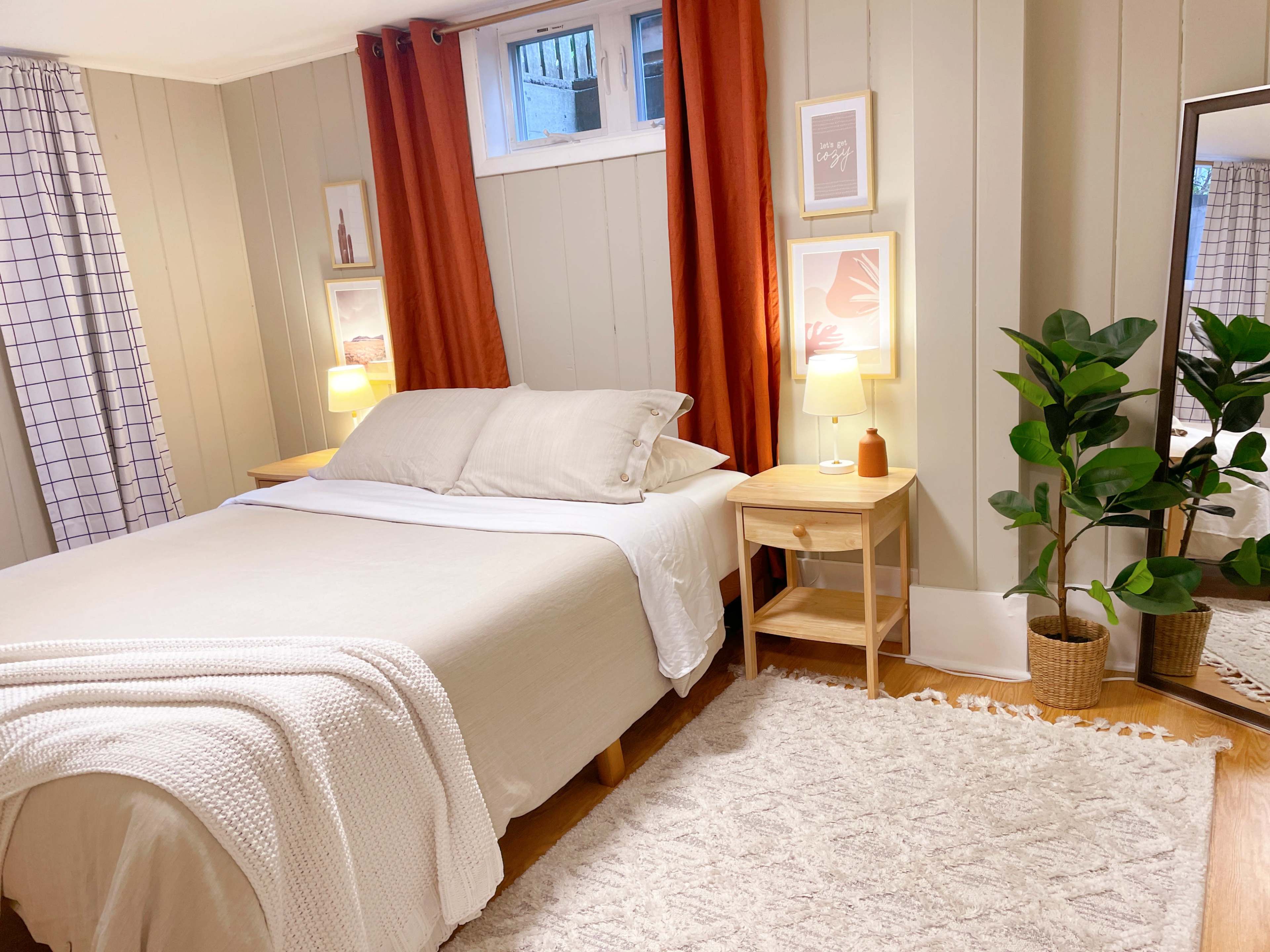 The image shows a cozy bedroom featuring a bed with neutral bedding, a pair of wooden nightstands with lamps, and decorative elements such as plants and framed artwork against light-colored walls.