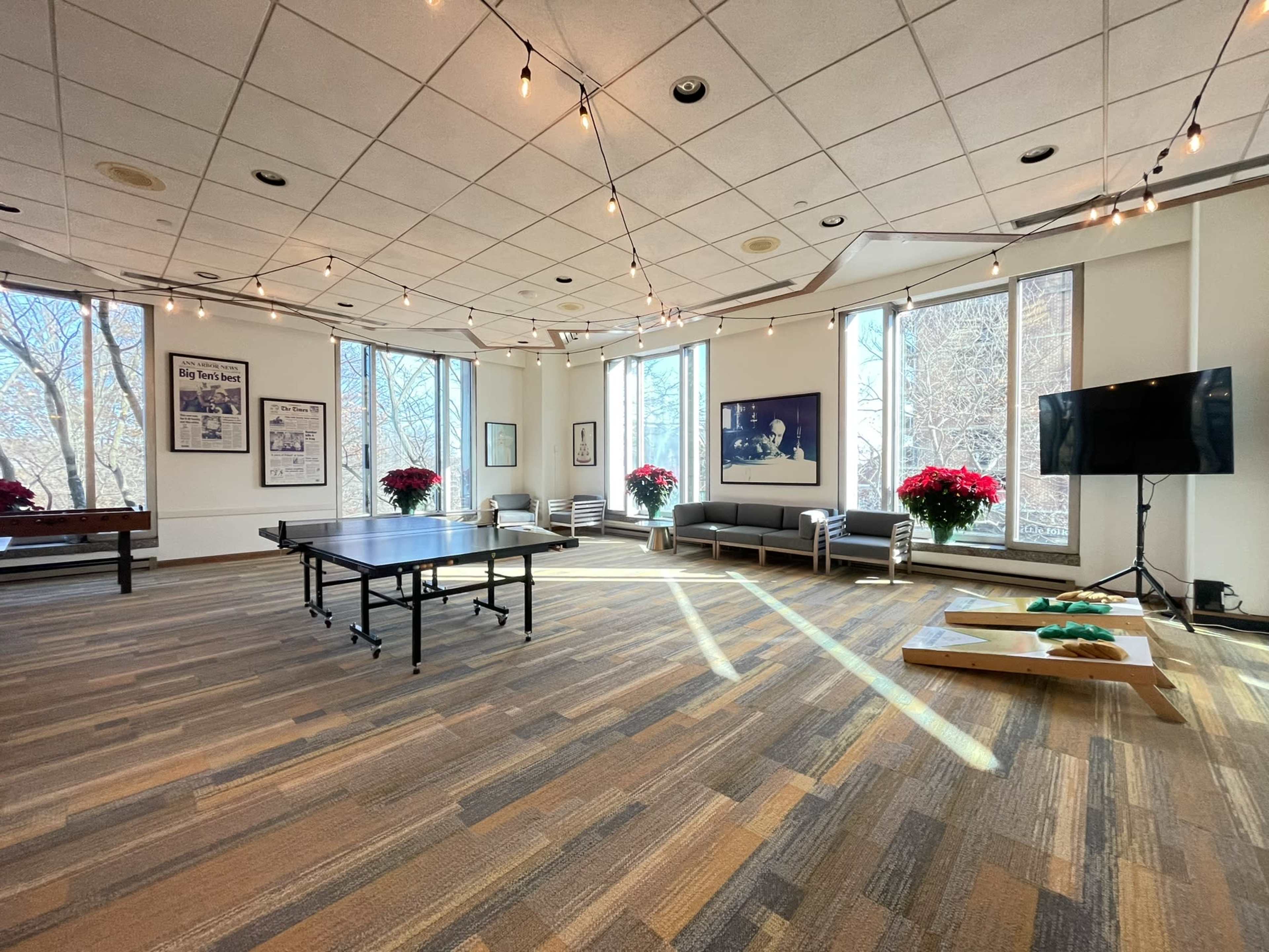 A spacious room with large windows, string lights overhead, a ping pong table, and a seating area adorned with potted plants.