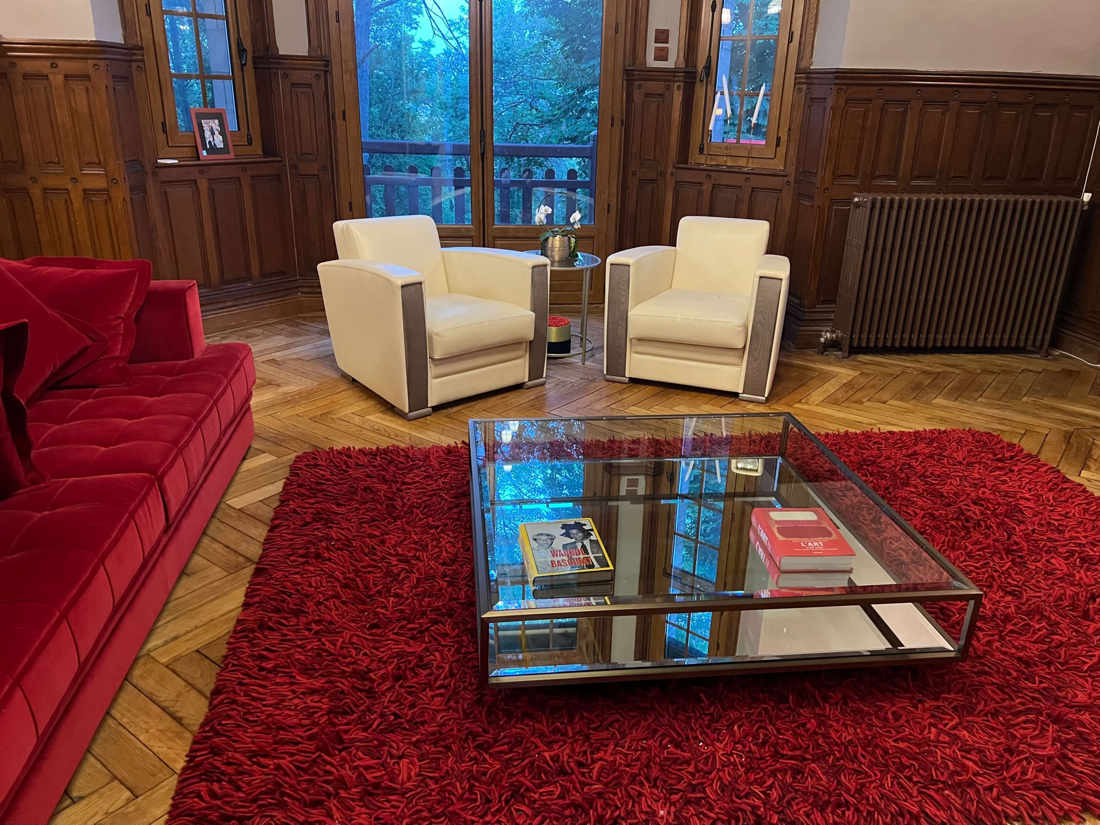 A living room with two white armchairs facing a glass coffee table on a red rug, surrounded by wood-paneled walls and a large window overlooking greenery.