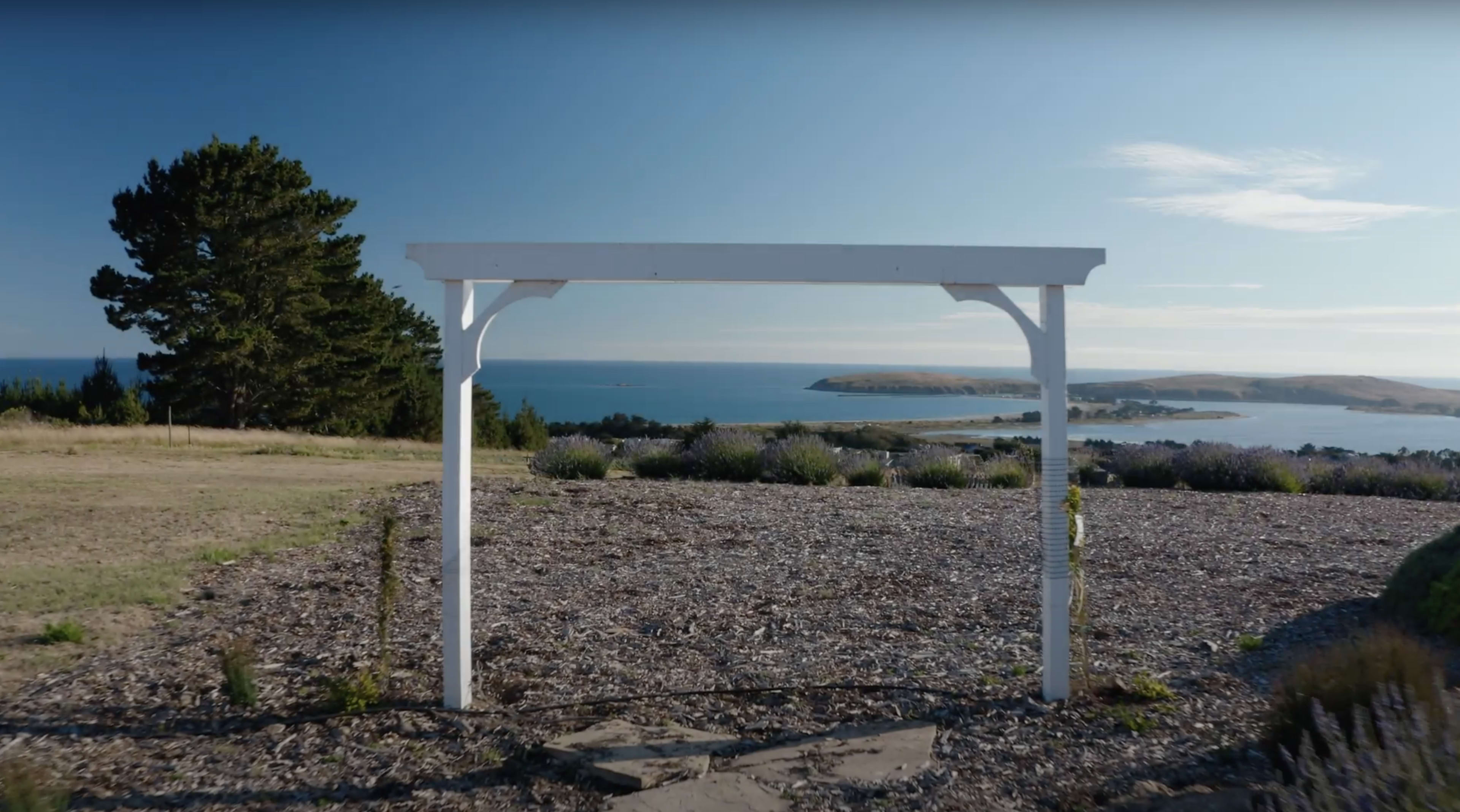 A white wooden structure stands on a gravel area overlooking a calm bay with distant hills.