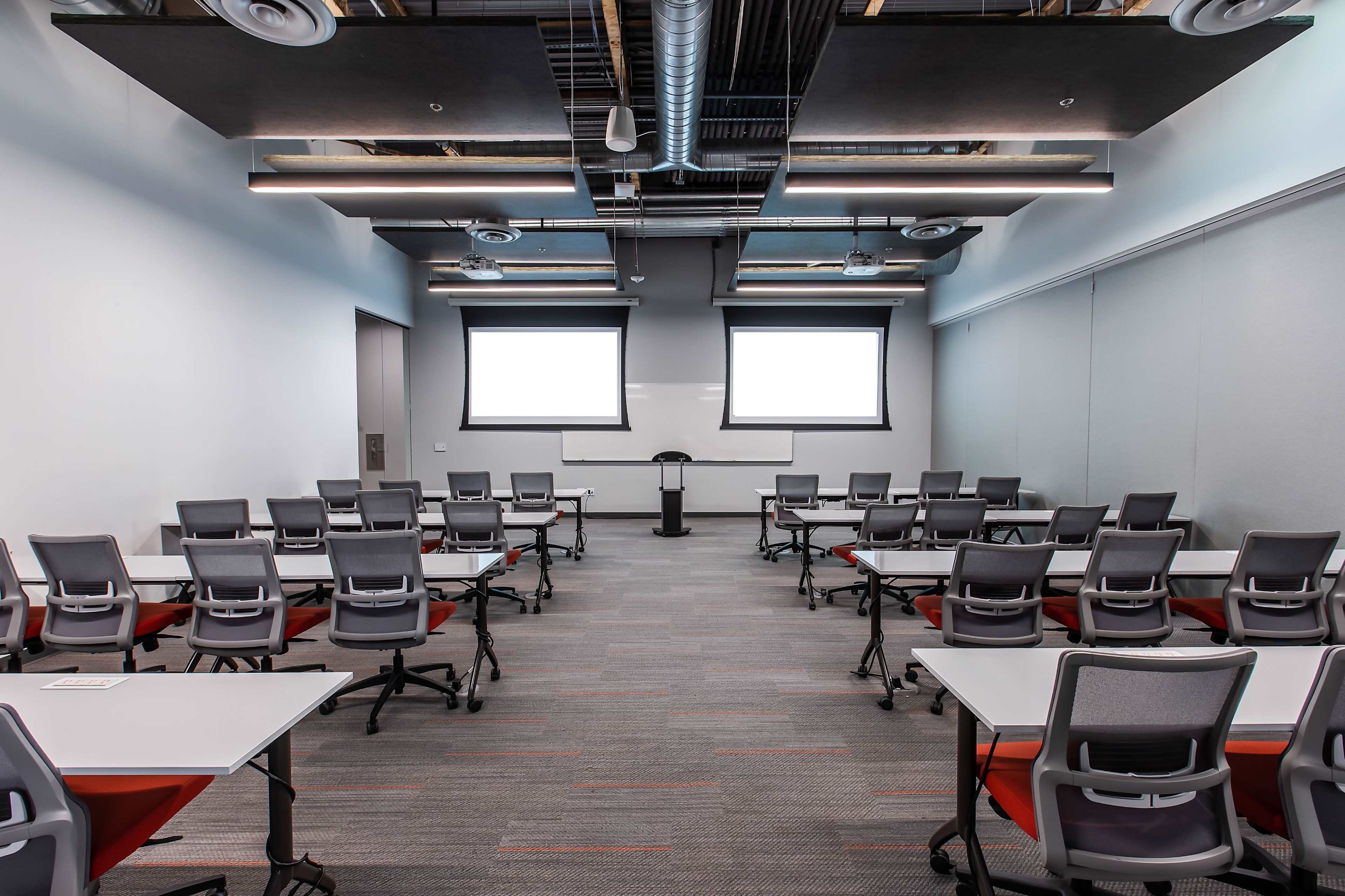 A training room setup with rows of tables and chairs facing a podium and two large projection screens.