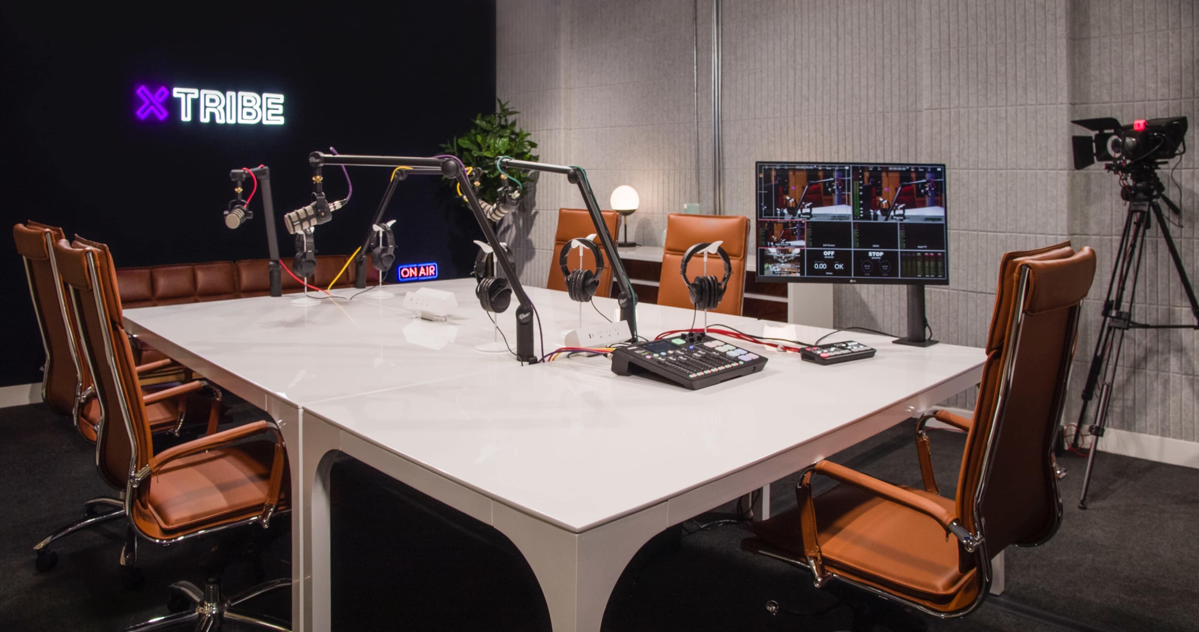 The image shows a modern podcasting studio with a white table, eight brown leather chairs, audio equipment, and multiple monitors displaying video feeds.