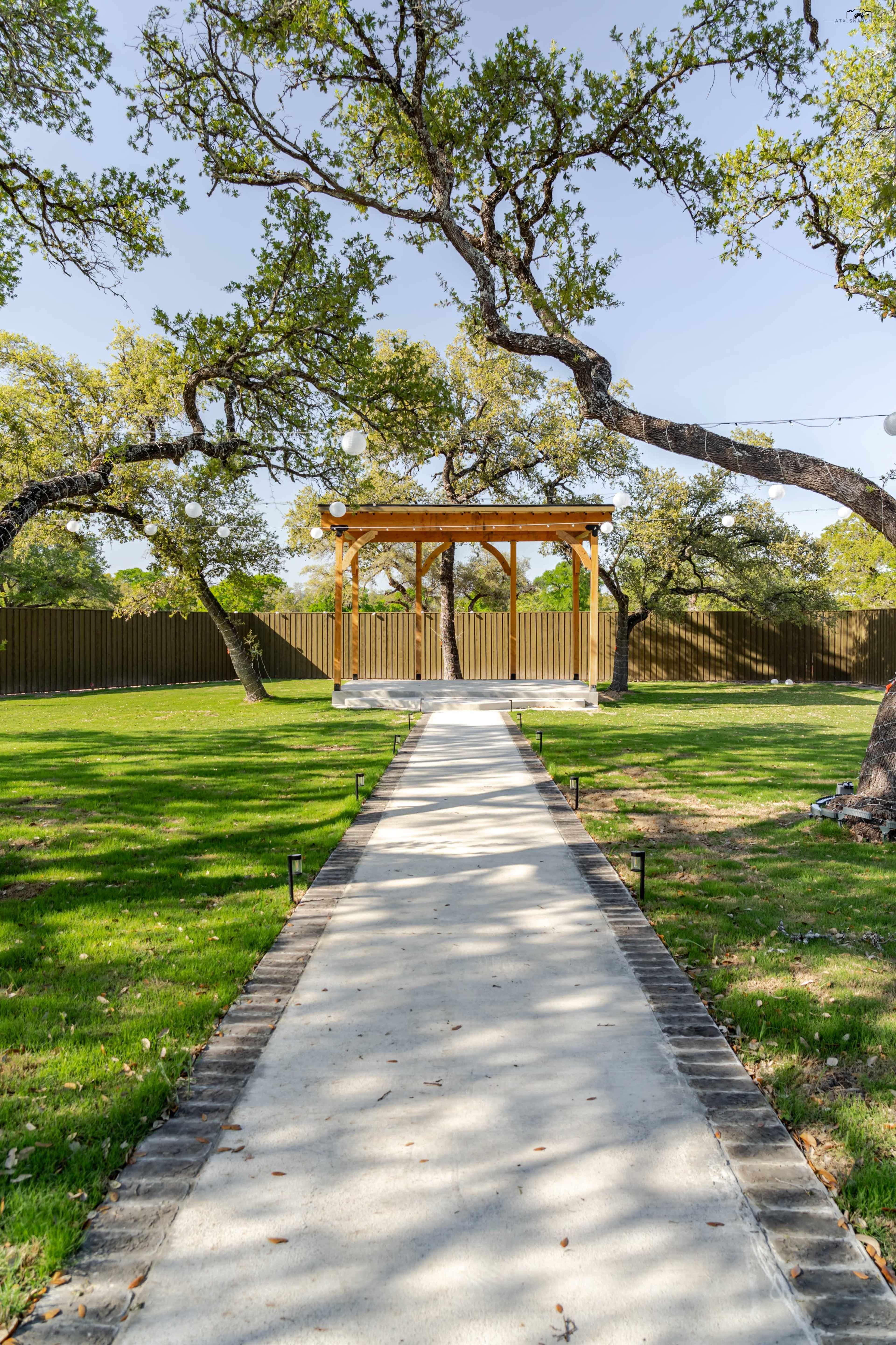 A concrete pathway leads to a wooden pavilion surrounded by trees in a green lawn.