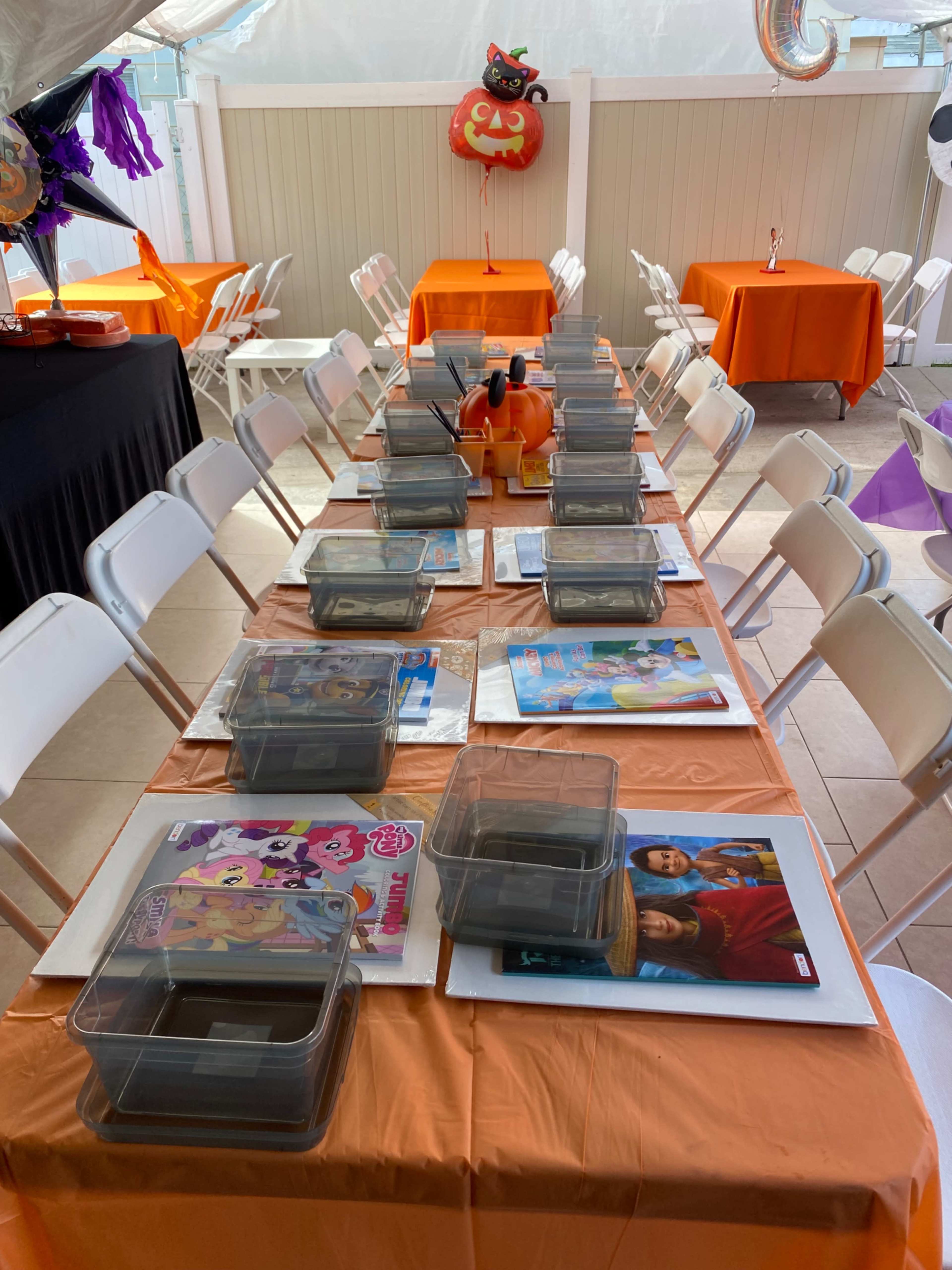 The image shows a party setup with orange and black tables arranged with clear containers and various colorful decorations in the background.