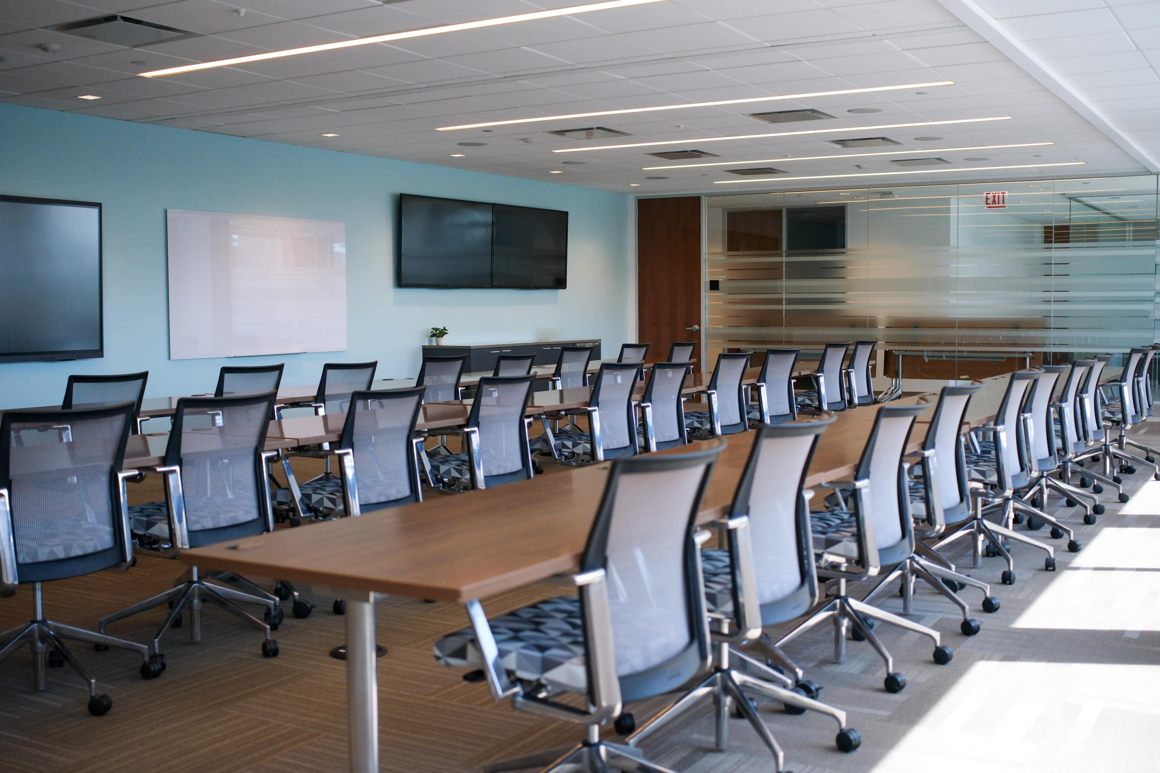 A modern conference room features multiple tables arranged in a U-shape with ergonomic chairs, large screens on the walls, and a light blue backdrop.