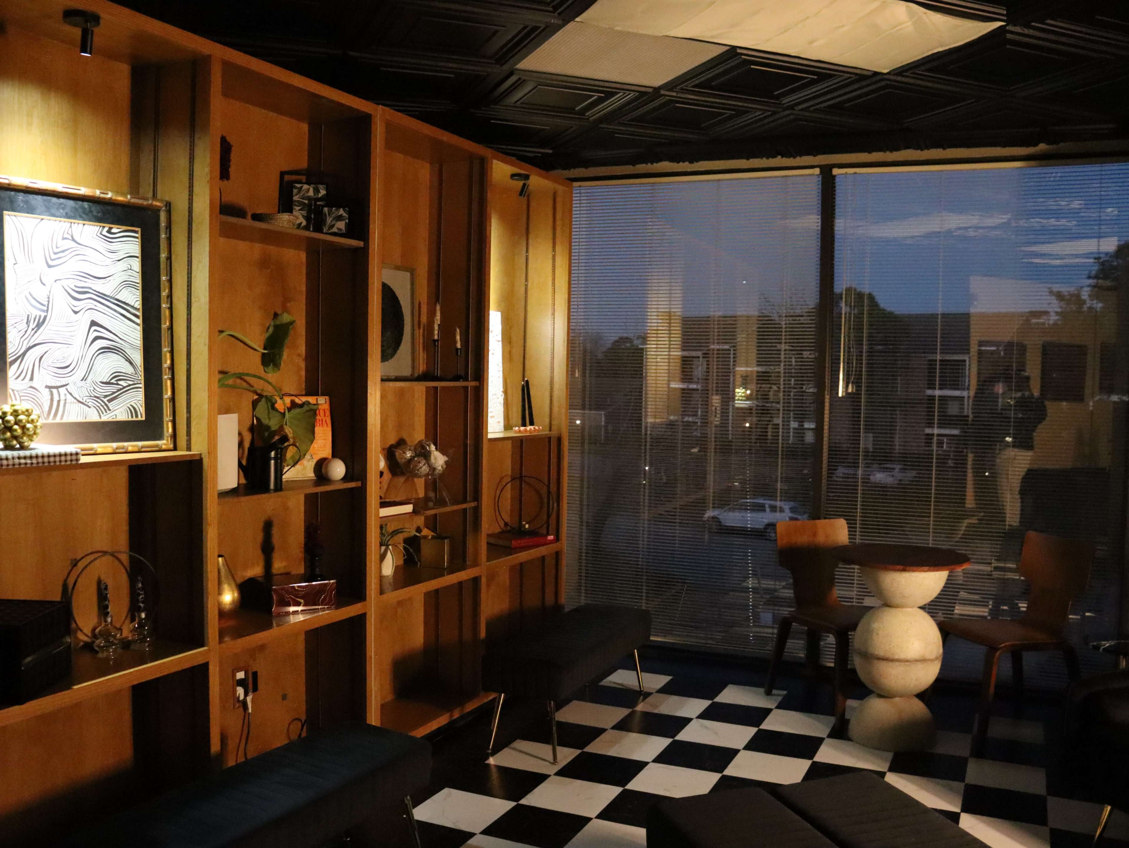 The image shows a modern interior with a checkered floor, wooden shelving displaying decorative items, and large windows partially covered by blinds, revealing a twilight view outside.