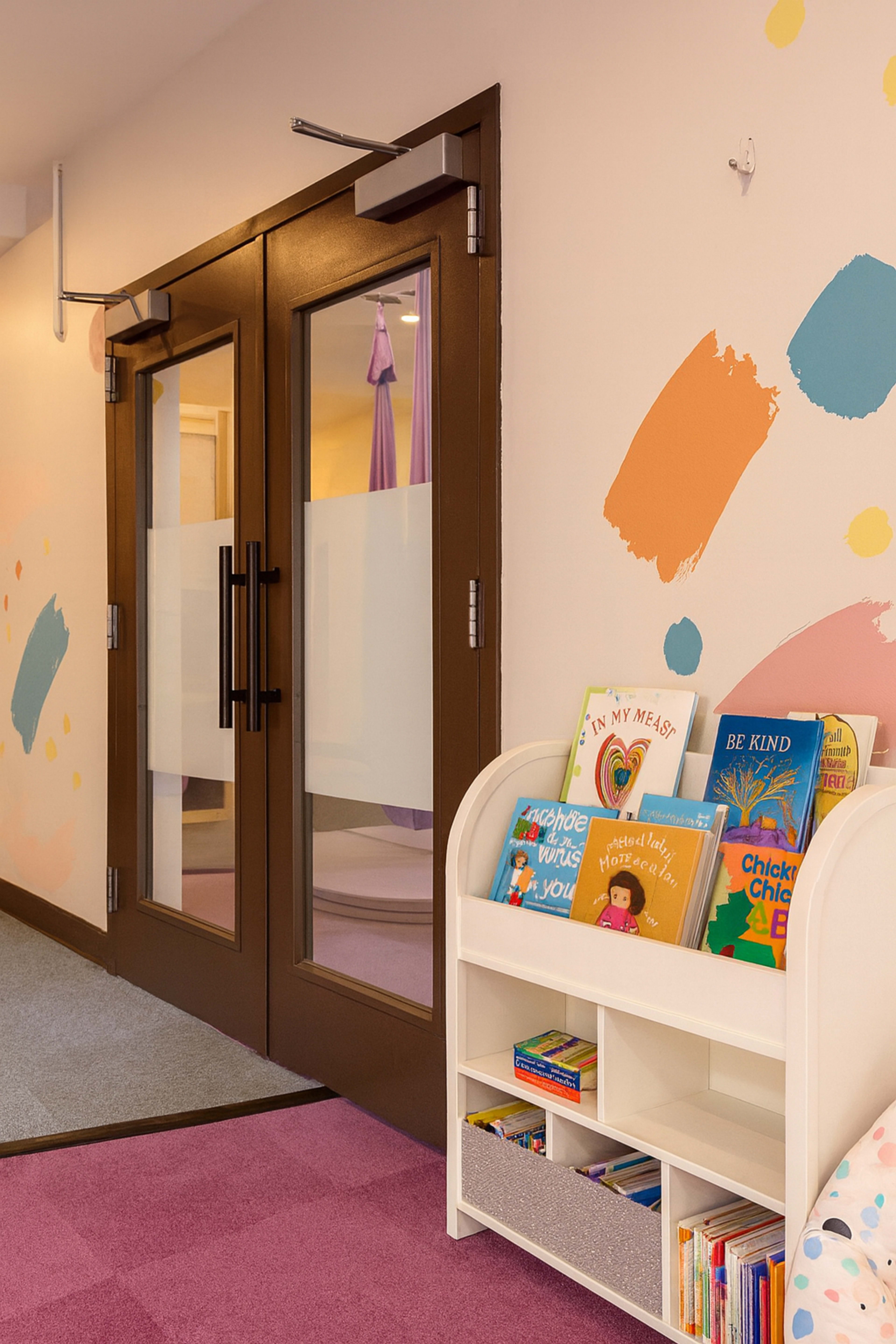 The image shows a colorful children's reading area with a white bookshelf filled with books and large double doors in the background.