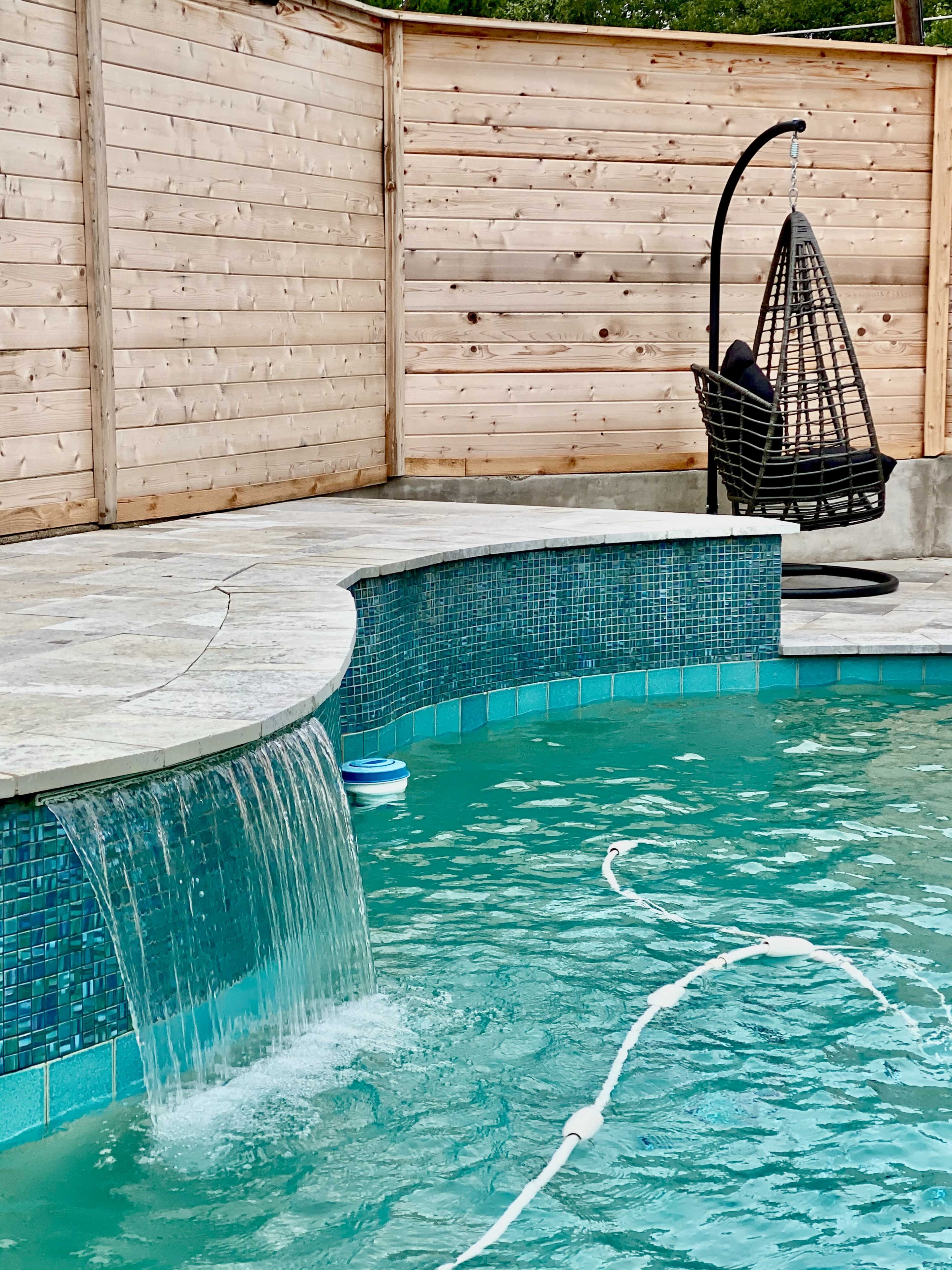 A swimming pool features a waterfall and a hanging chair beside a wooden fence.