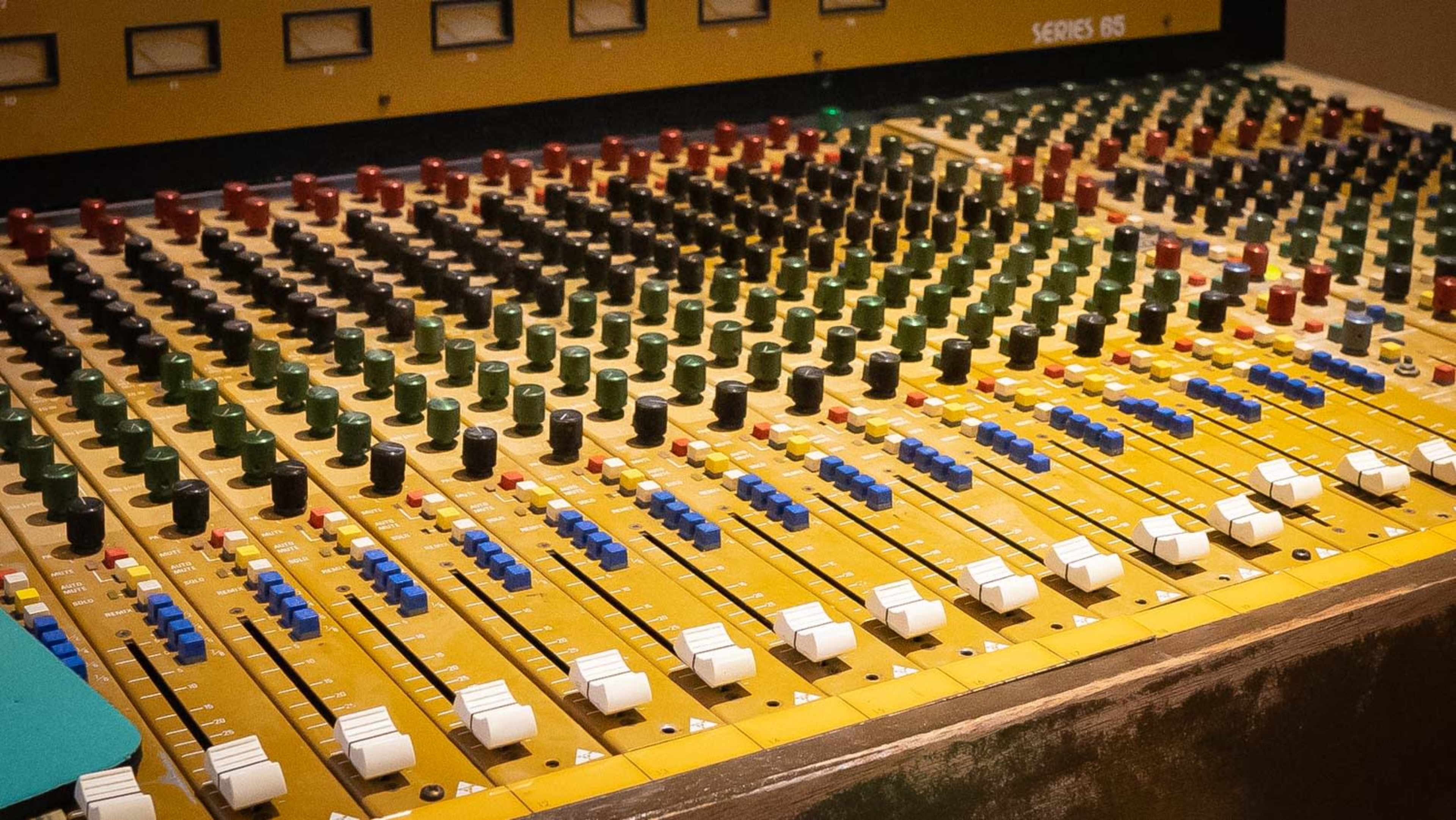 A close-up of a sound mixing console with various colored faders and knobs arranged on a yellow surface.