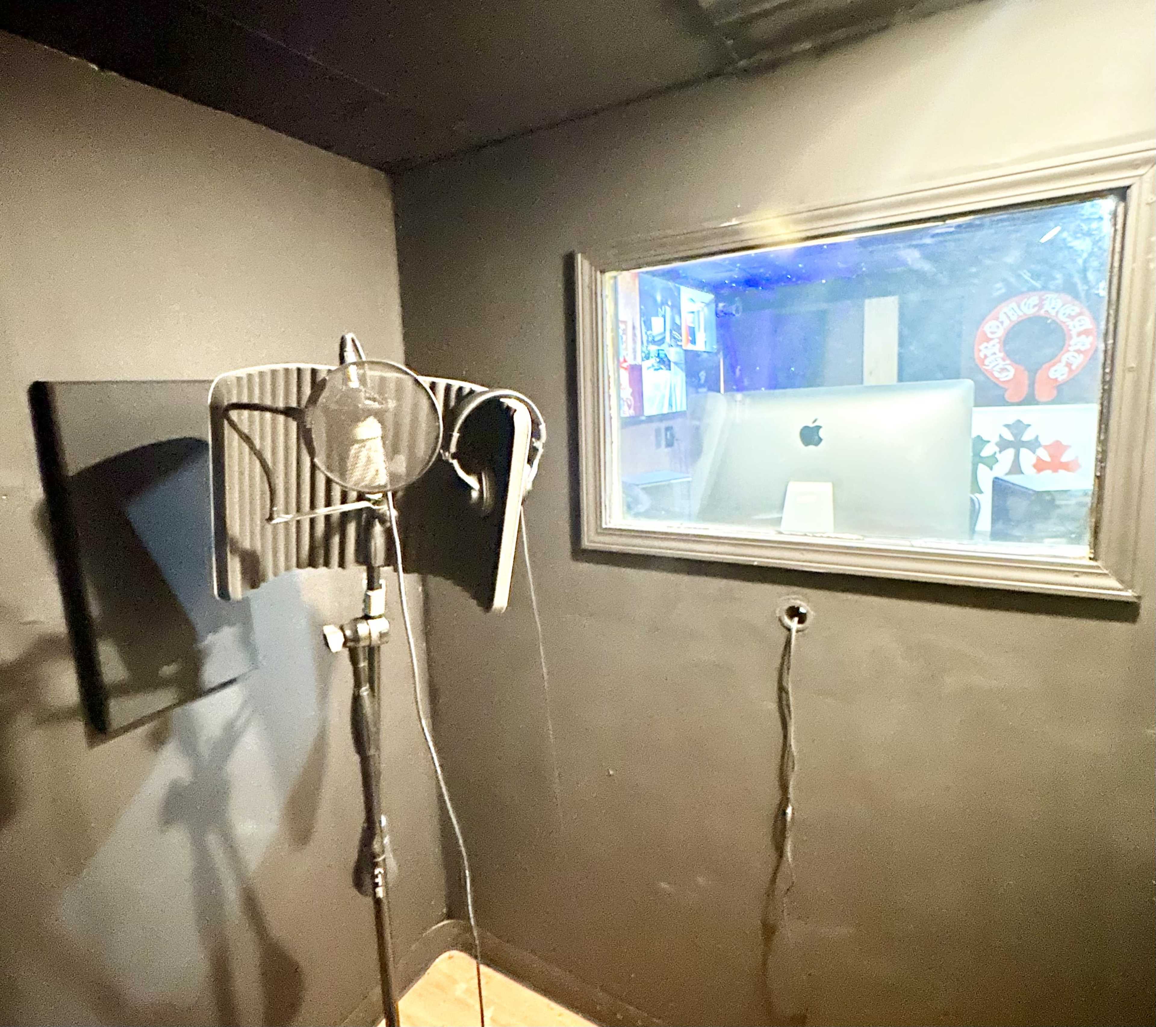 The image shows a small vocal recording booth with a microphone on a stand and a window viewing into another room containing an Apple computer.