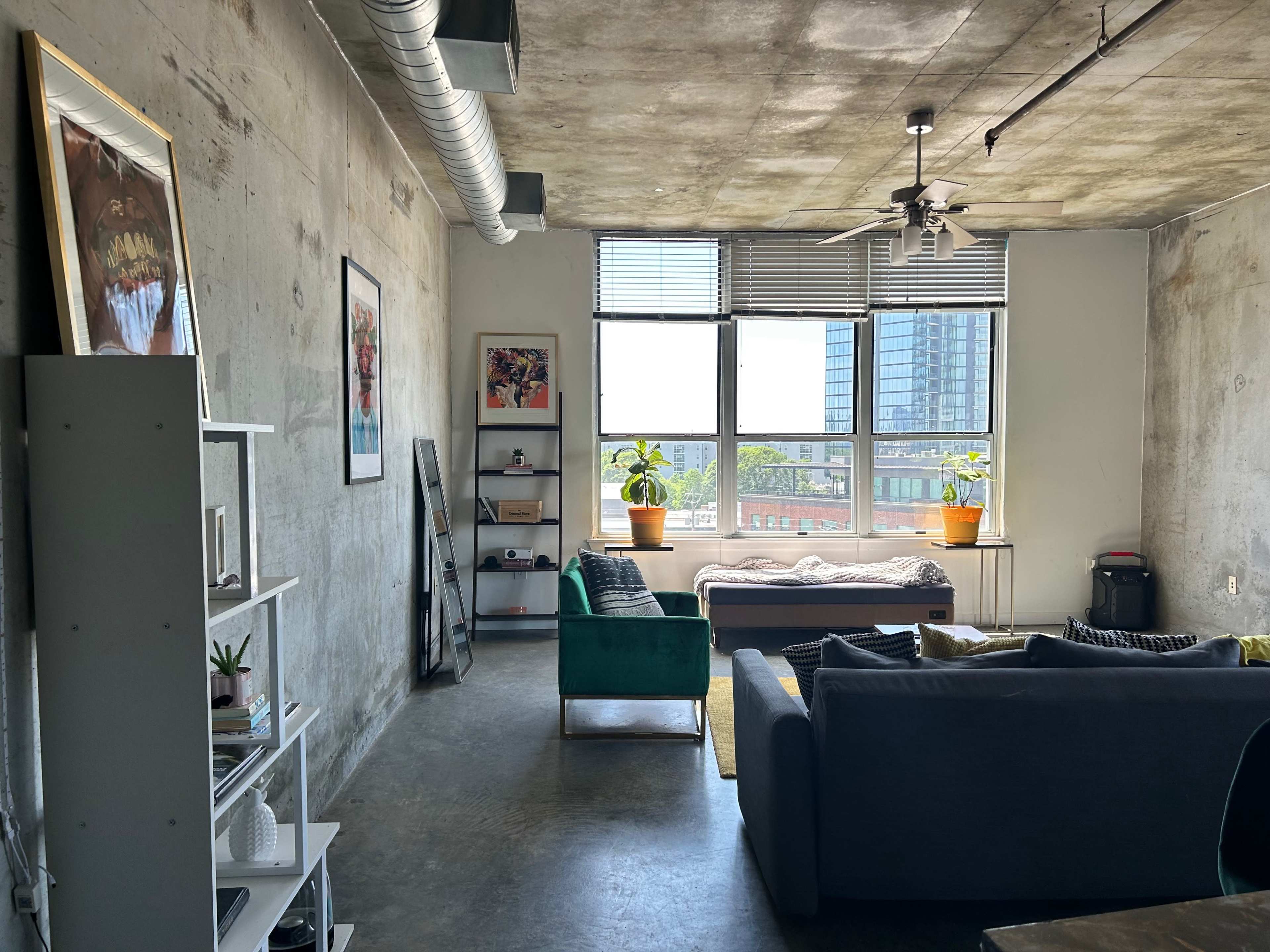 The image shows a modern apartment with concrete walls, large windows, and a green sofa facing a well-lit area with a bed and indoor plants.