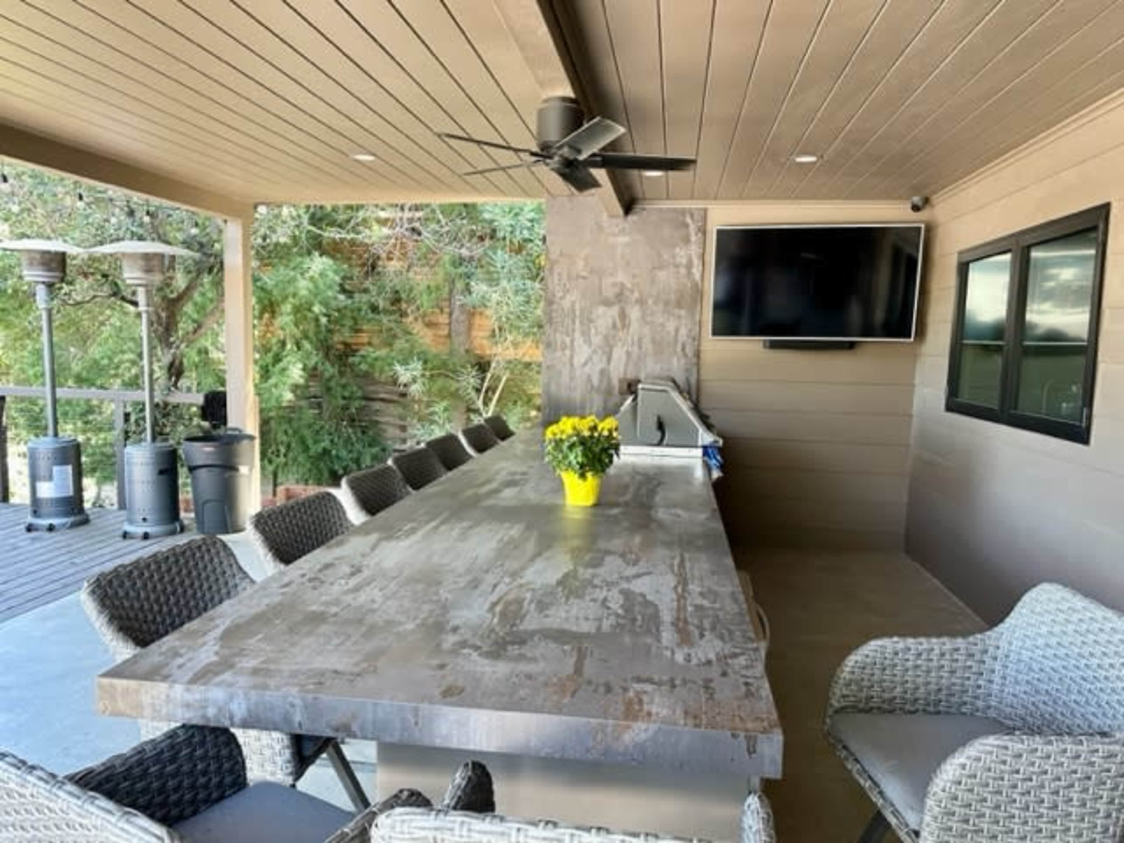 A covered outdoor patio with a large stone table, a yellow flower centerpiece, a television mounted on the wall, and several chairs arranged around the table.