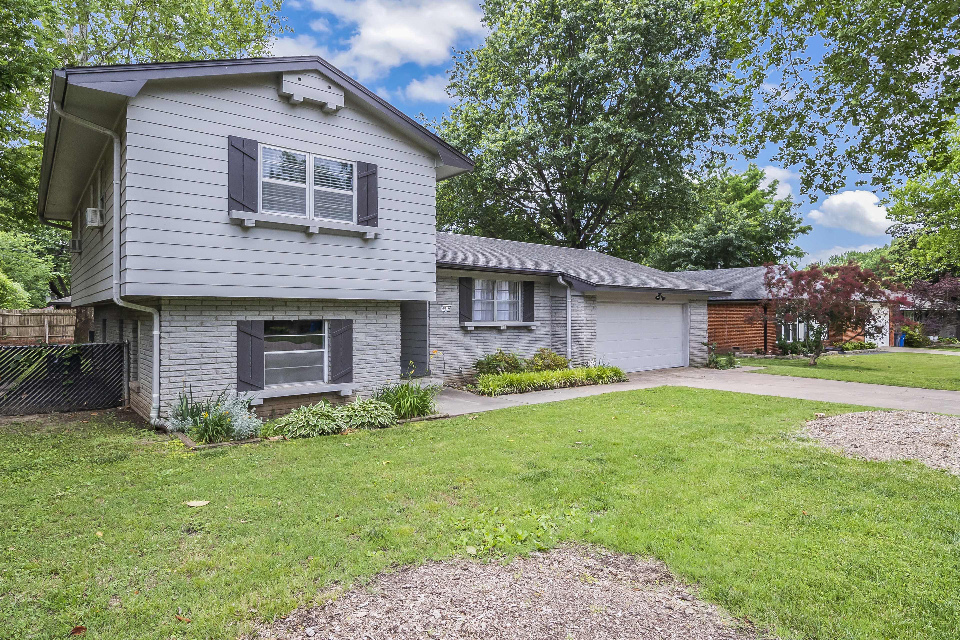 The image shows a two-story gray house with a brick foundation, surrounded by a grassy yard and several trees.