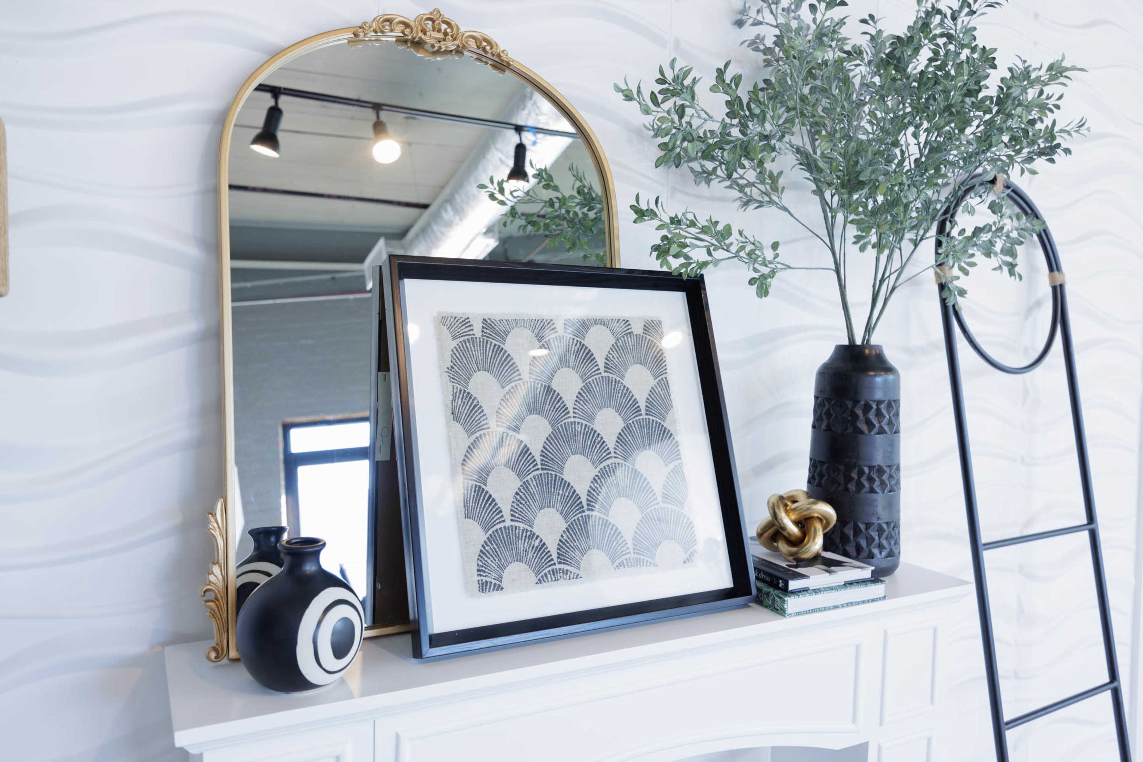 A decorative table features a framed art piece, a potted plant, a black vase, and a small stack of books, all set against a mirrored backdrop.