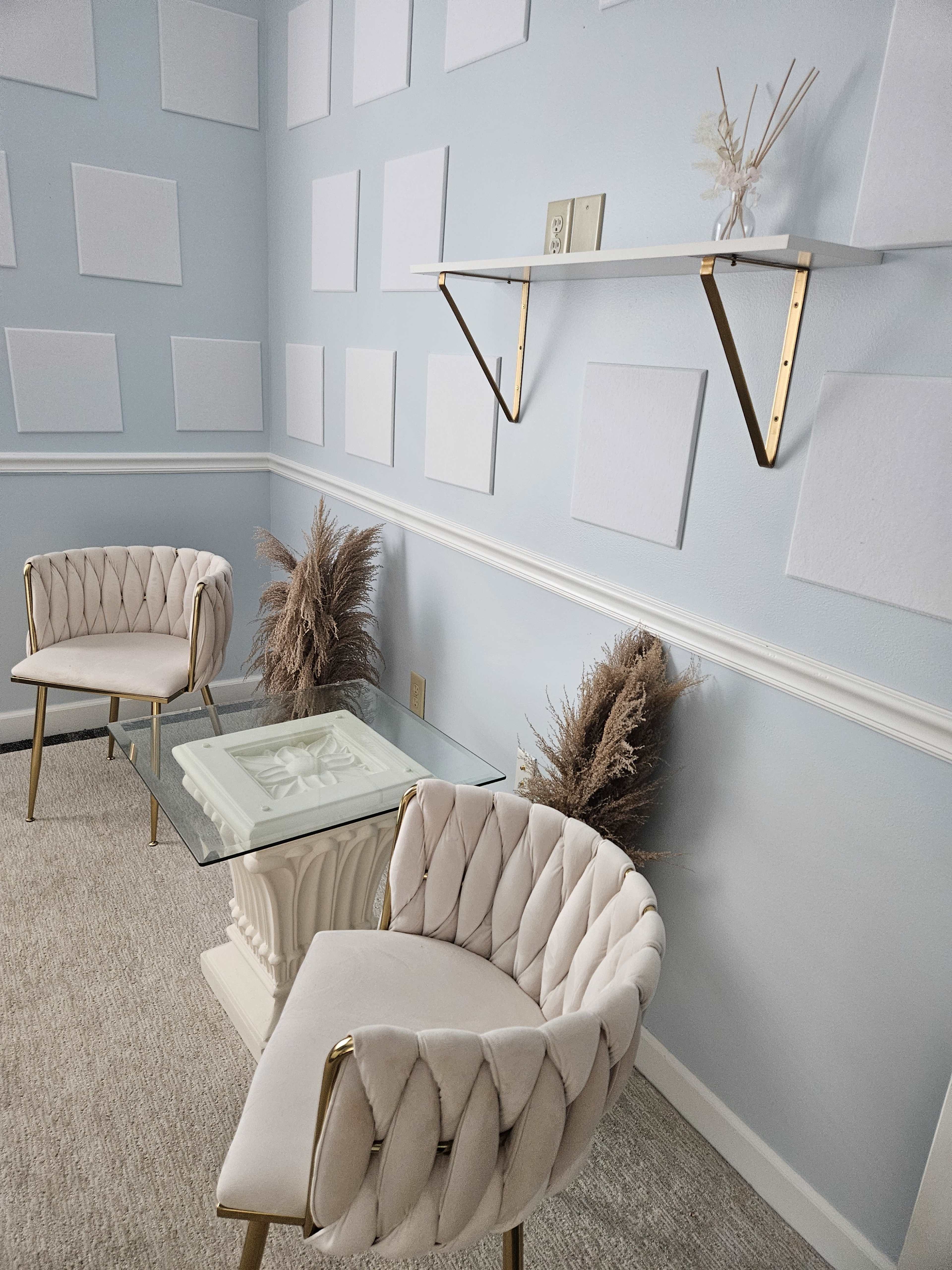 The image shows a light blue room with two beige upholstered chairs, a glass table with a decorative base, and a shelf above featuring white square panels on the wall.