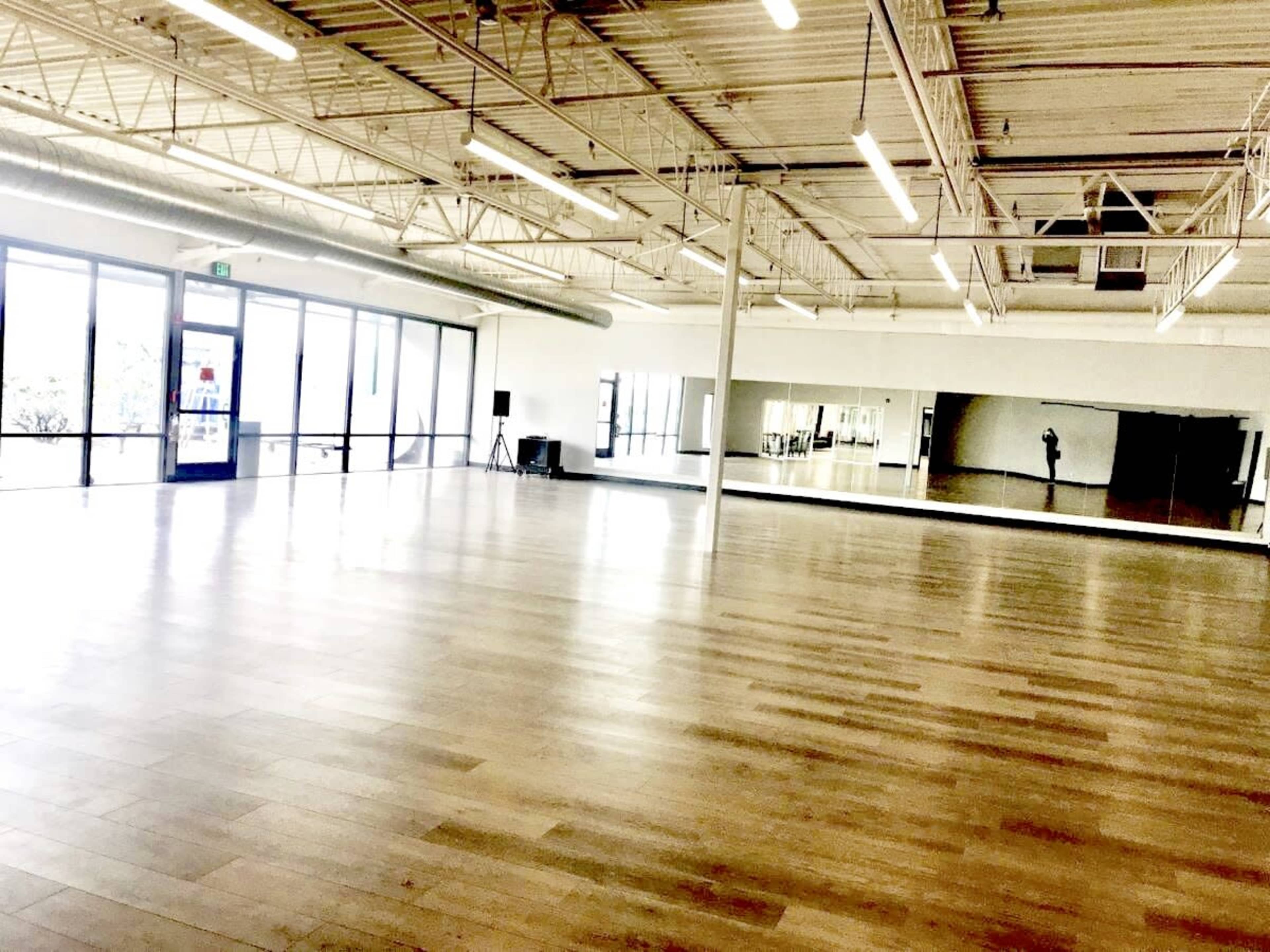 An empty dance studio with large windows, a mirrored wall, and wooden flooring.