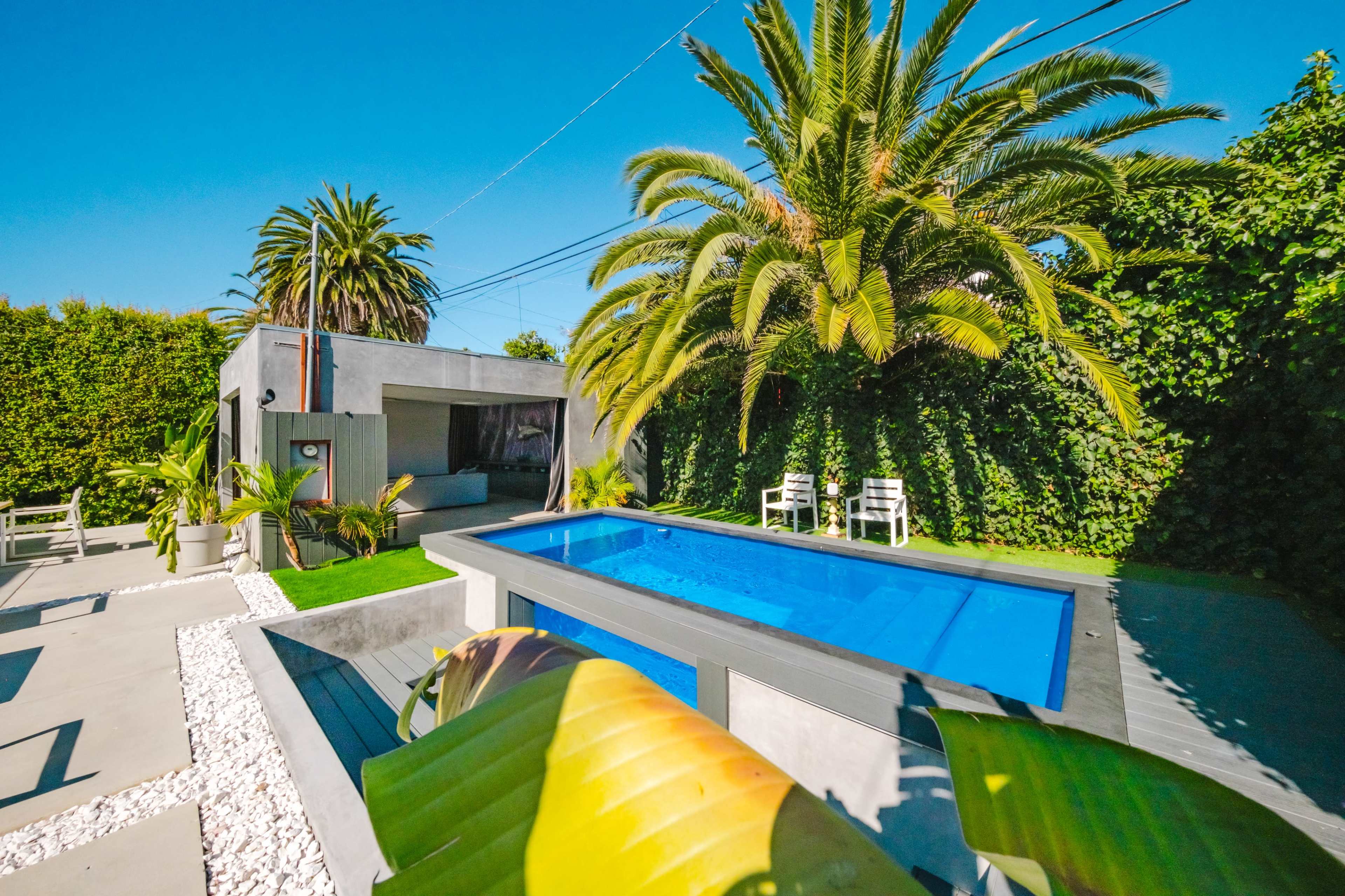The image features a modern backyard with a swimming pool surrounded by palm trees and a small seating area.