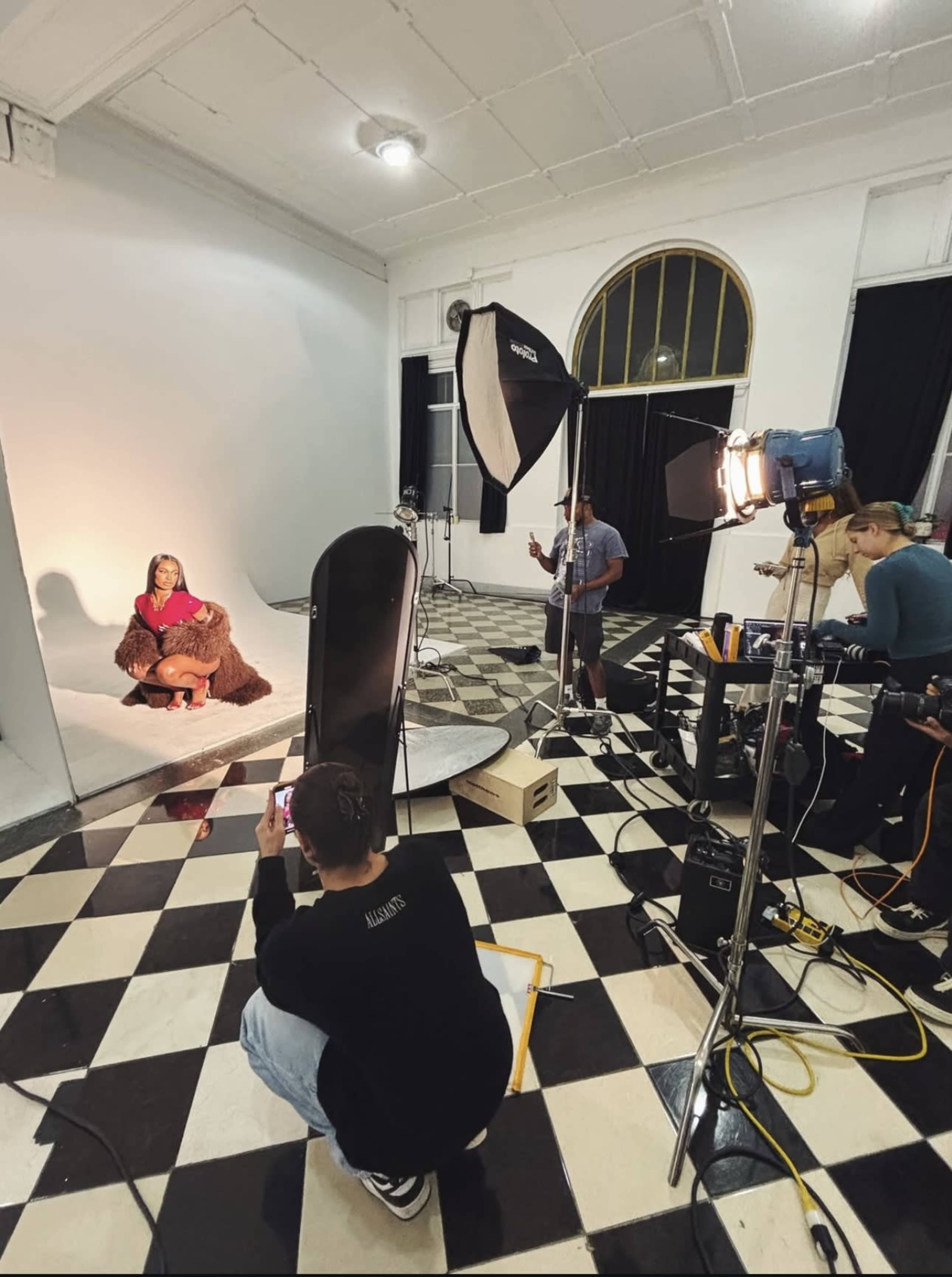A photoshoot is taking place in a studio with a model in a brown outfit, surrounded by equipment and photographers.