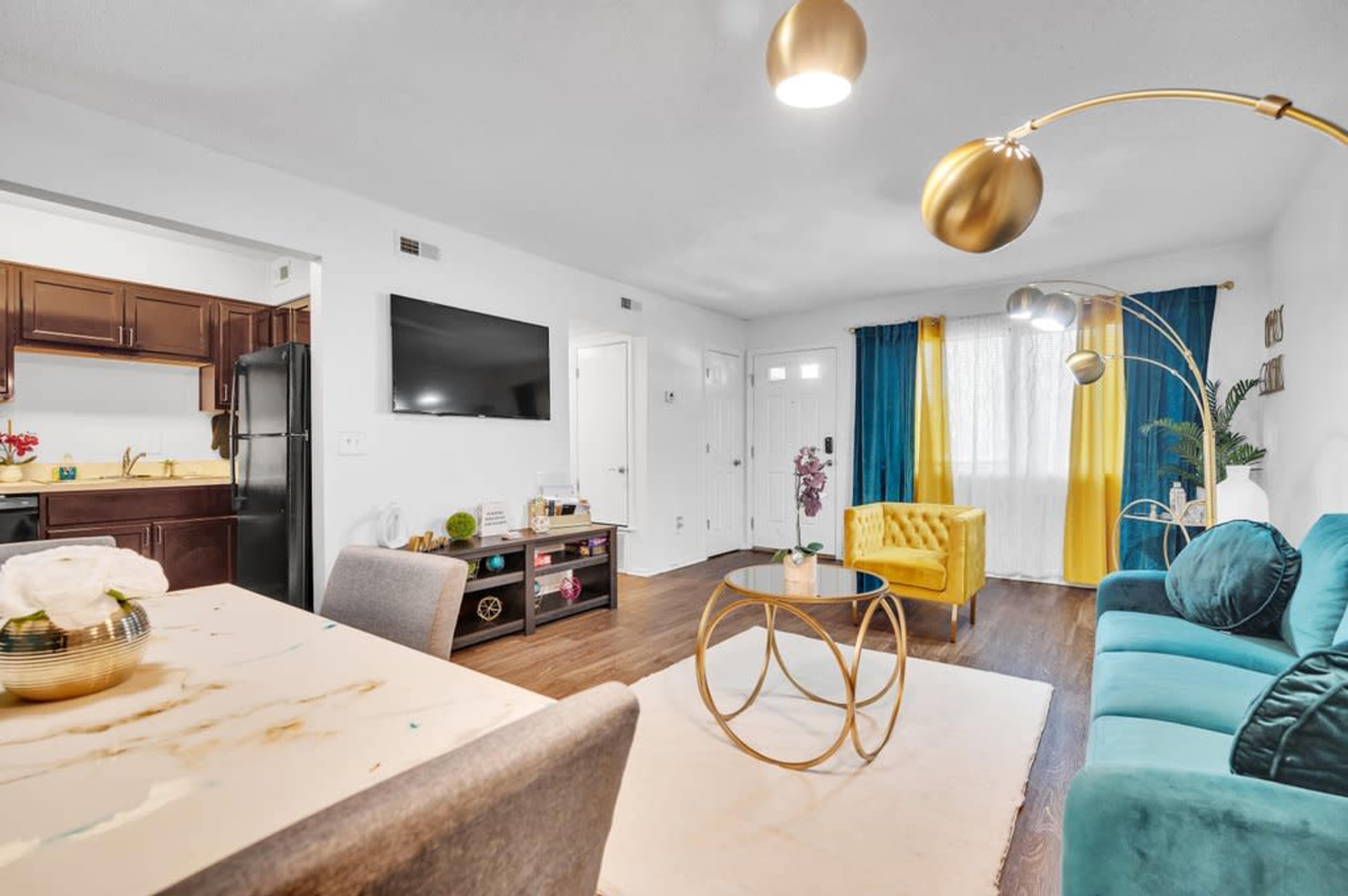 A well-lit living room features a teal sofa, a yellow armchair, and a round coffee table, with a television mounted on the wall and curtains in blue and yellow.