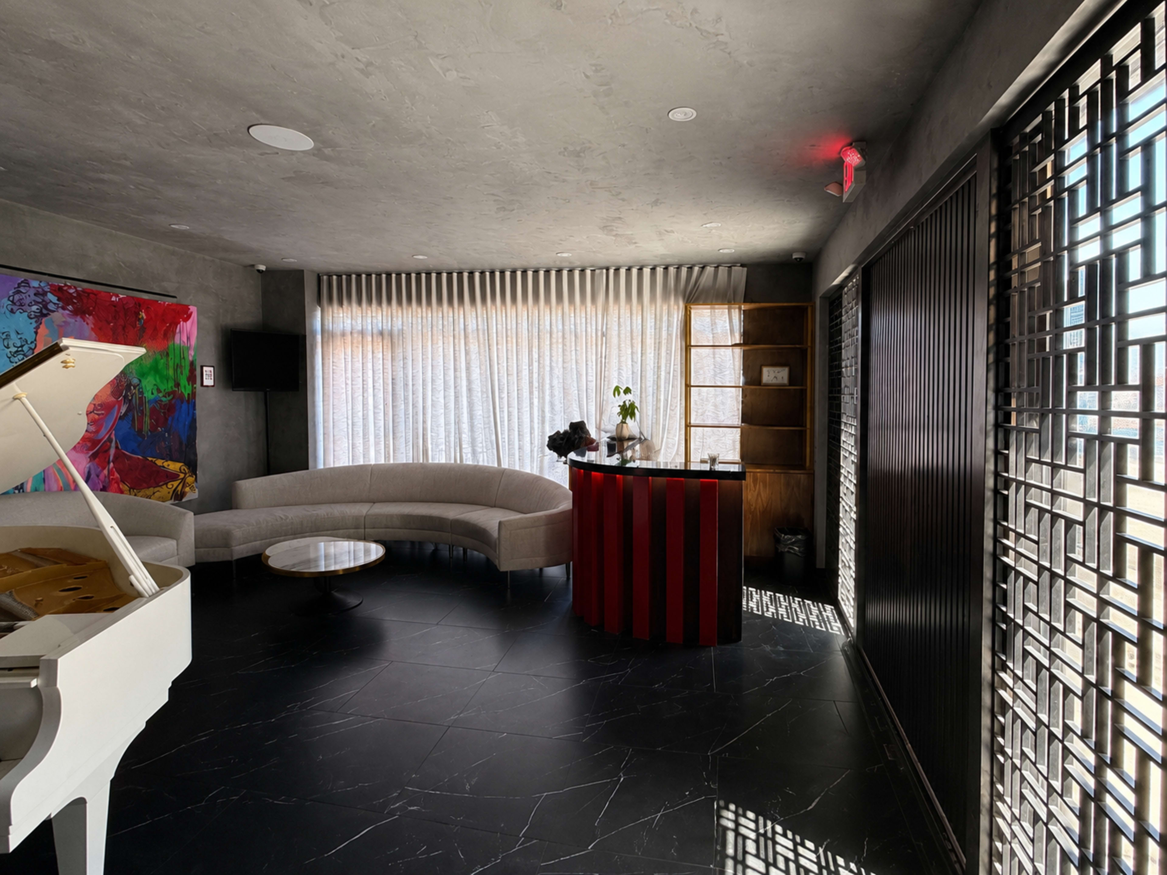 The image shows a modern interior space featuring a curved sofa, a circular coffee table, a piano, and a red-striped reception desk, with large windows covered by sheer curtains.