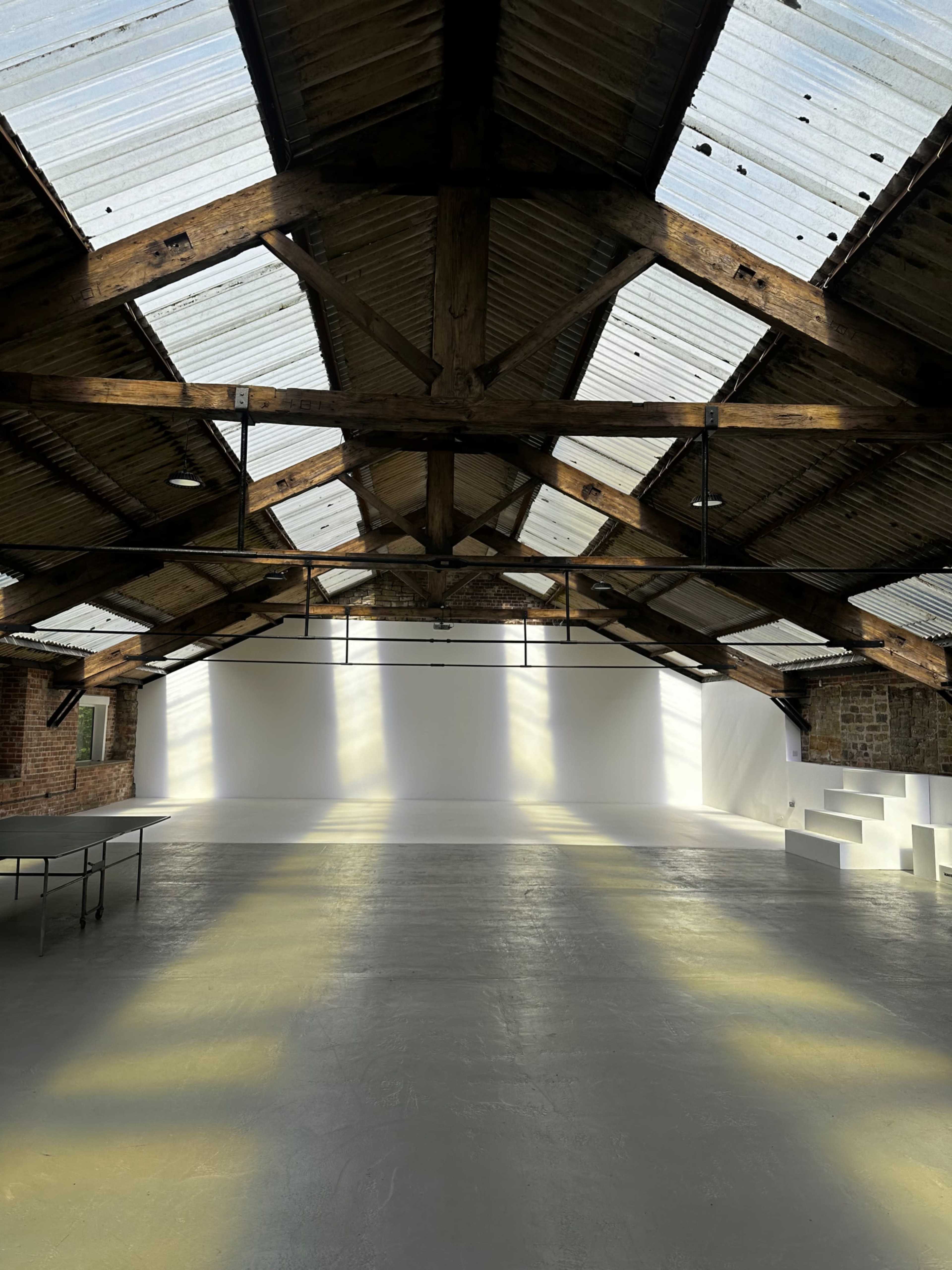 The image shows the interior of a spacious warehouse with a wooden truss ceiling, large windows, and a blank white wall at one end.