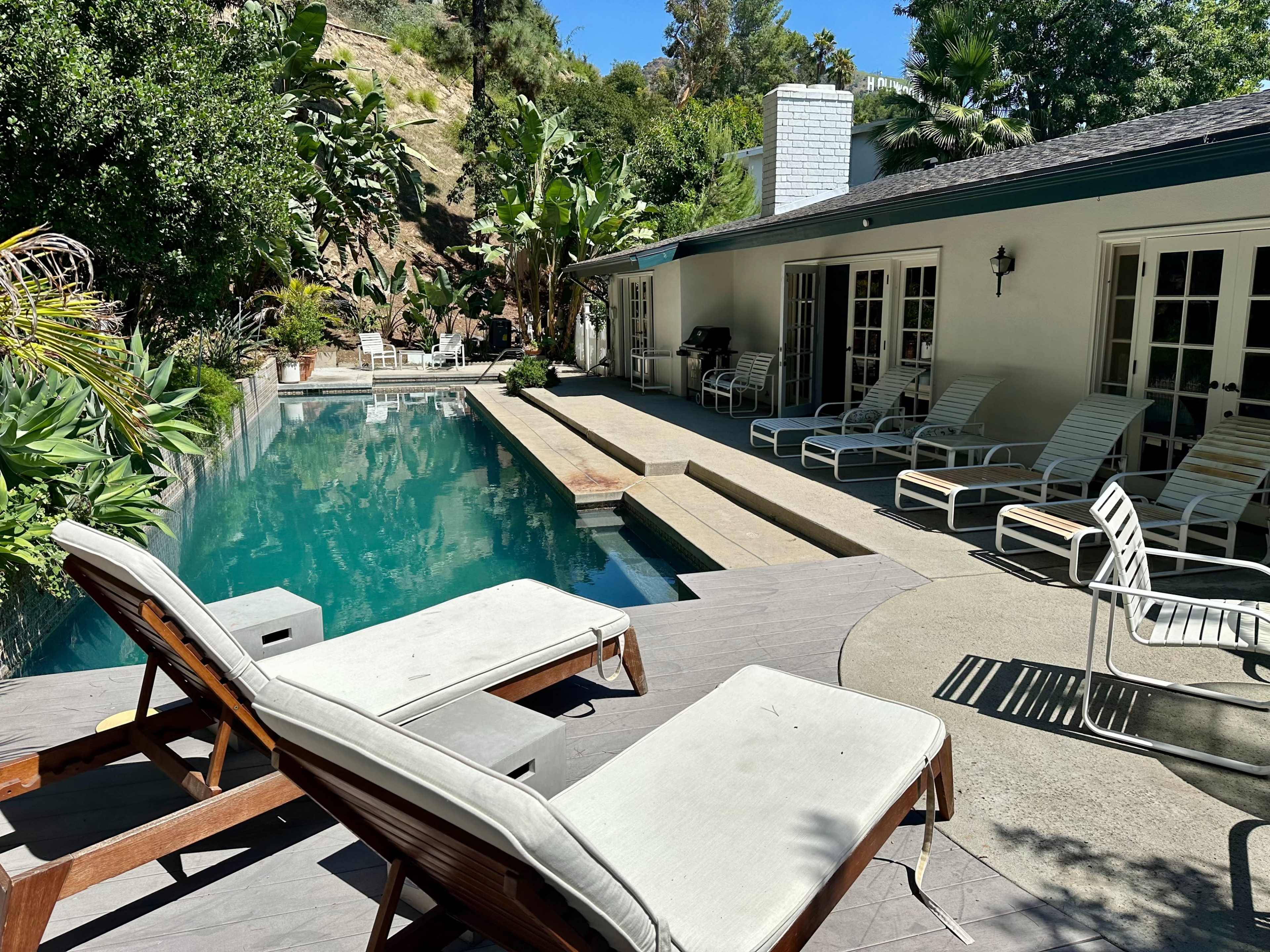 The image shows a backyard pool area with lounge chairs and steps leading to the water, surrounded by plants and a house in the background.