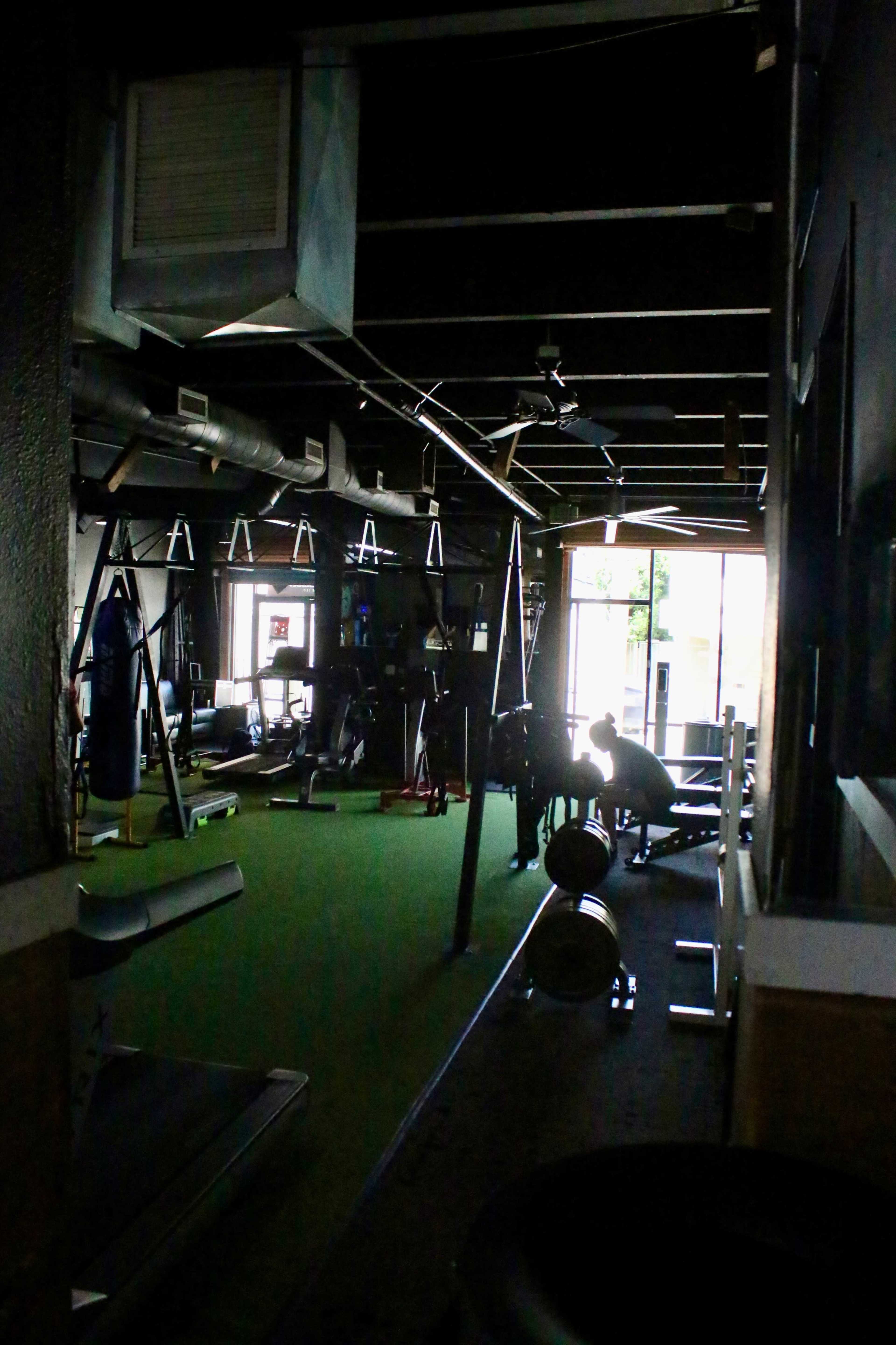 A dimly lit gym features exercise equipment and a person lifting weights near the entrance.