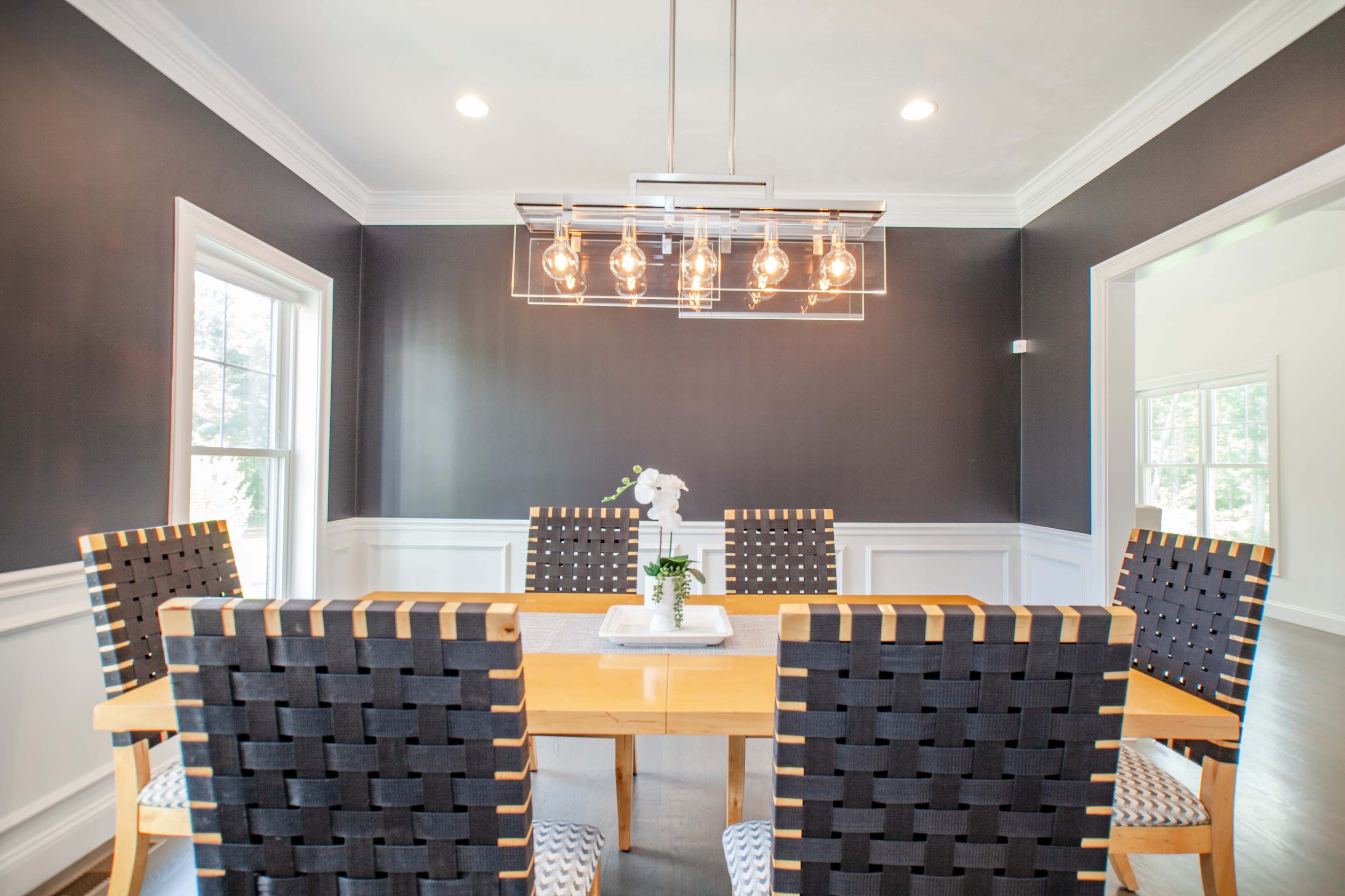 A modern dining room features a wooden table surrounded by dark woven chairs, with a stylish light fixture above and large windows providing natural light.