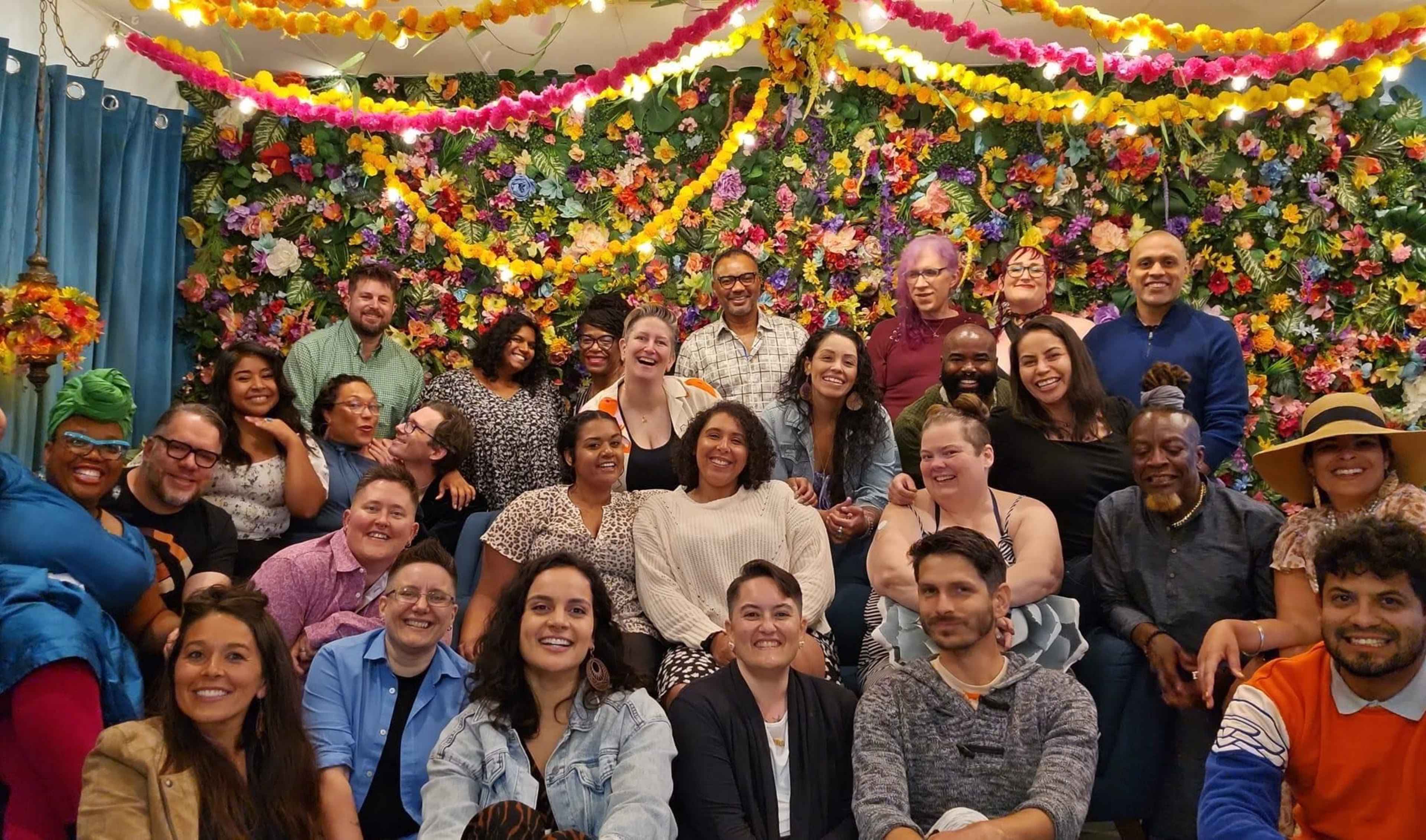 A diverse group of people is gathered in front of a colorful floral backdrop, smiling and posing for a photo.