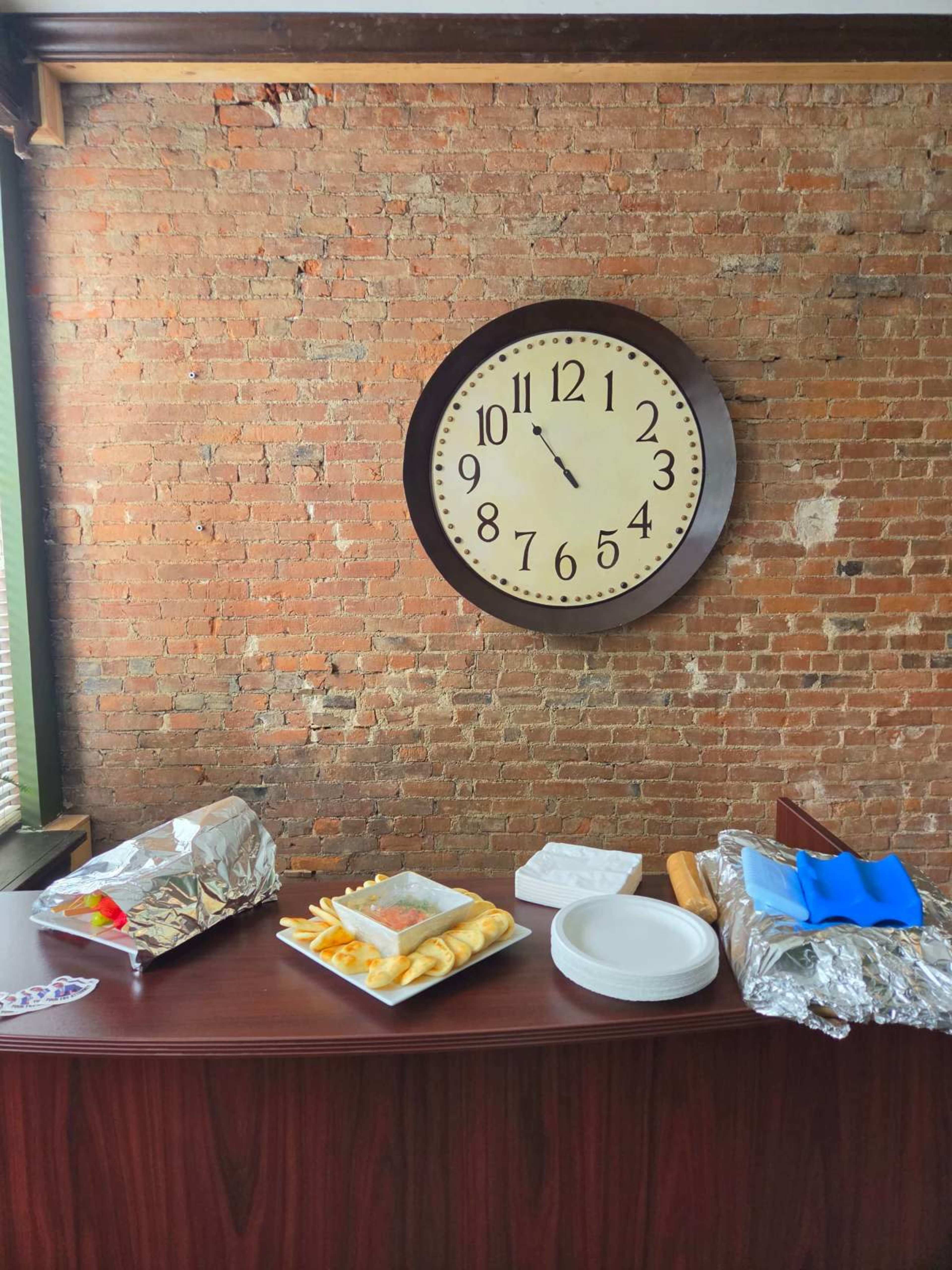 A large wall clock hangs above a table set with food items, including trays of snacks and disposable plates, against a brick wall.