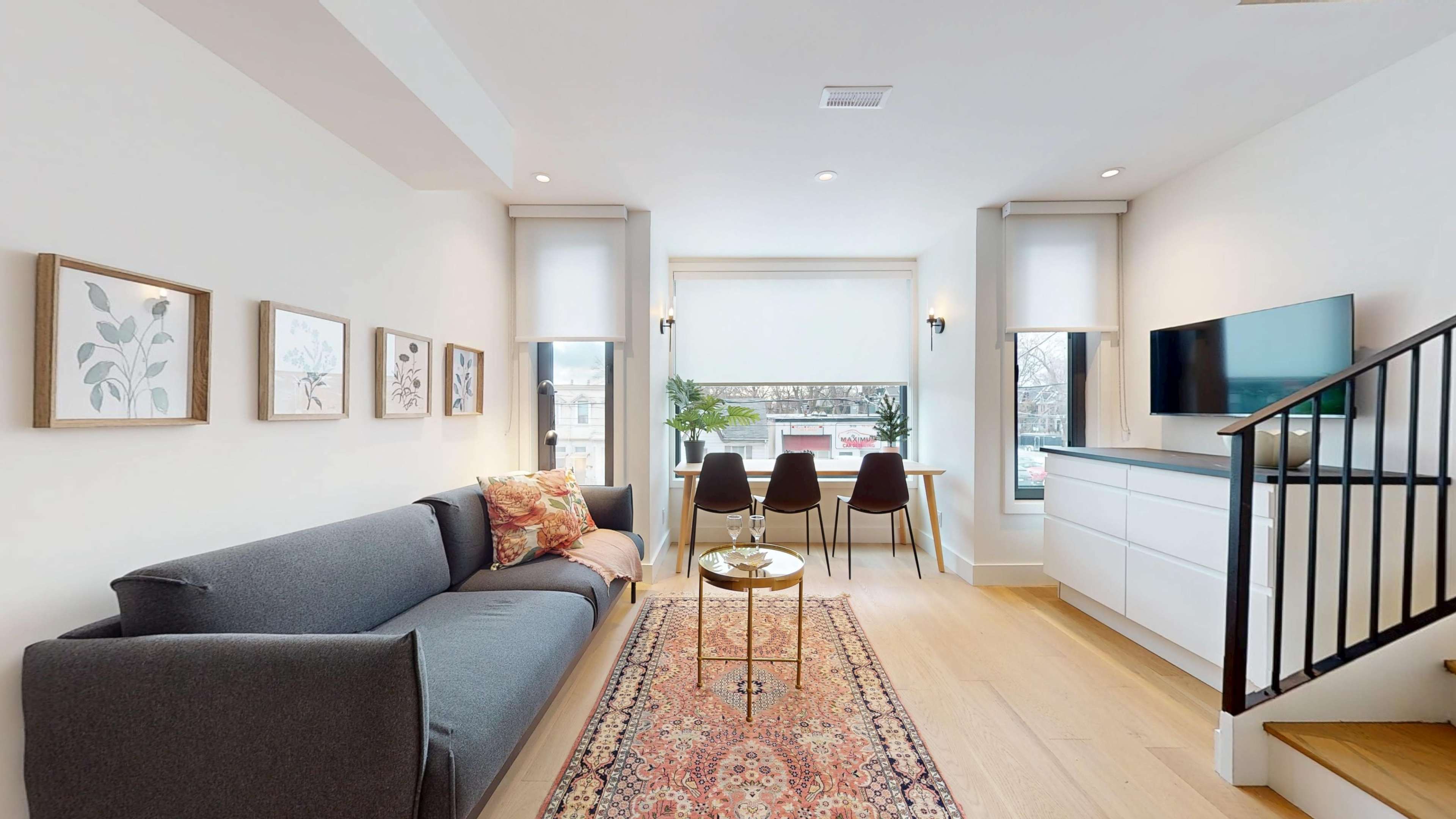 The image shows a modern living room with a gray sofa, a small round table, a dining area with four chairs, and framed art on the walls.