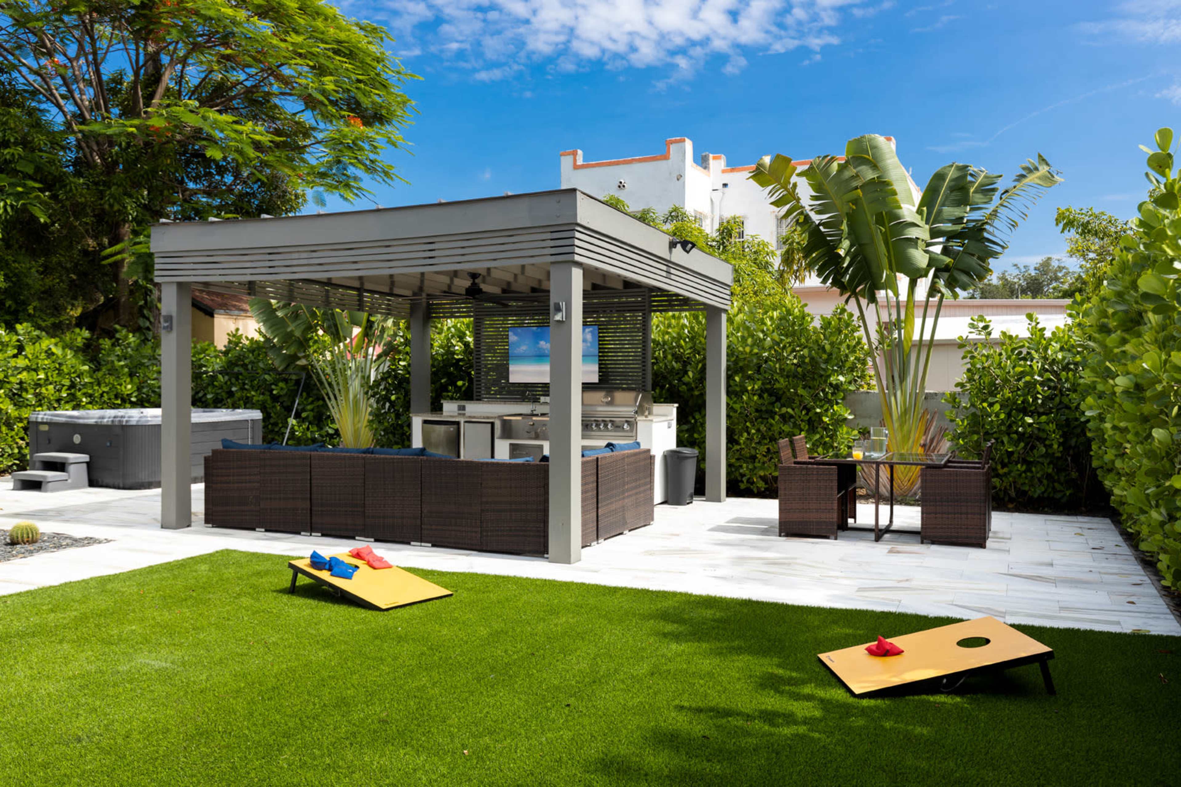 The image shows a backyard patio with a covered seating area, a grill, and cornhole boards on a grassy lawn.