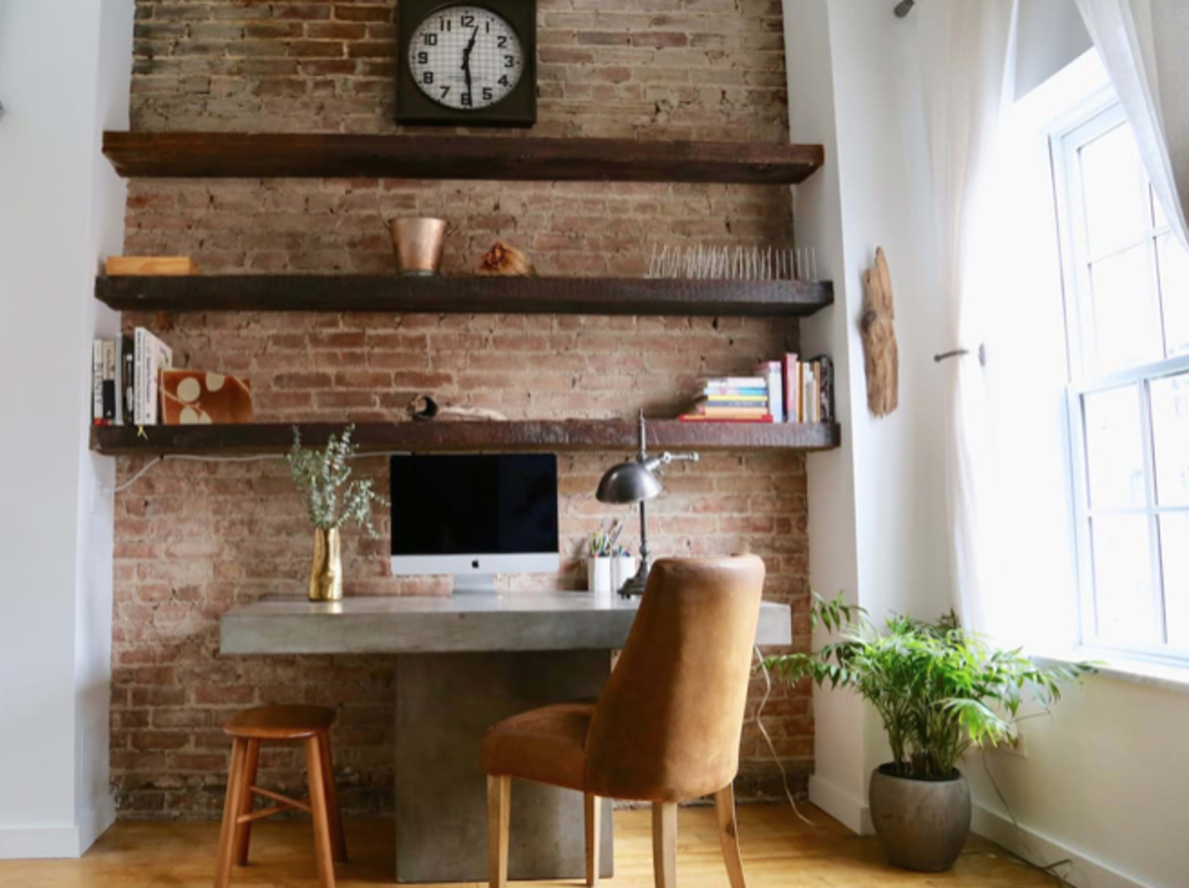 A modern workspace with a concrete desk, a computer, wooden shelves with books and decor, and a chair next to a potted plant in front of a window.