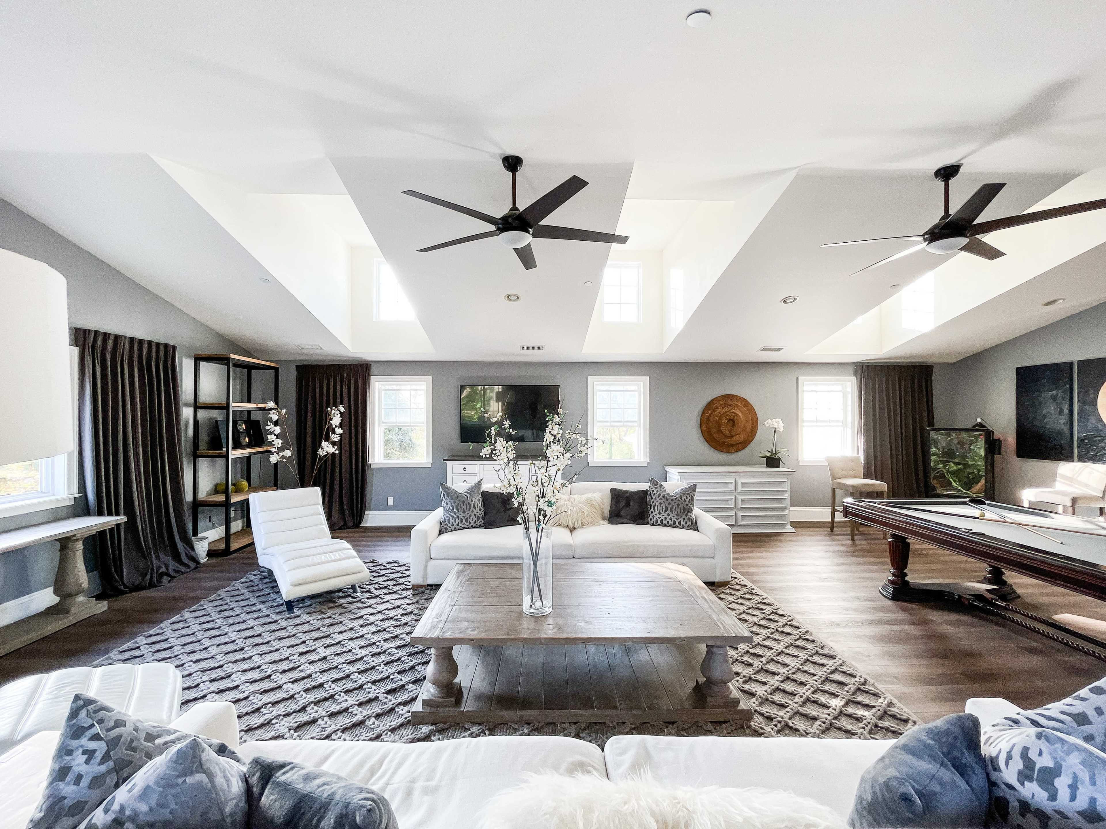 The image shows a spacious living room featuring two ceiling fans, large windows, light-colored sofas, and a pool table.