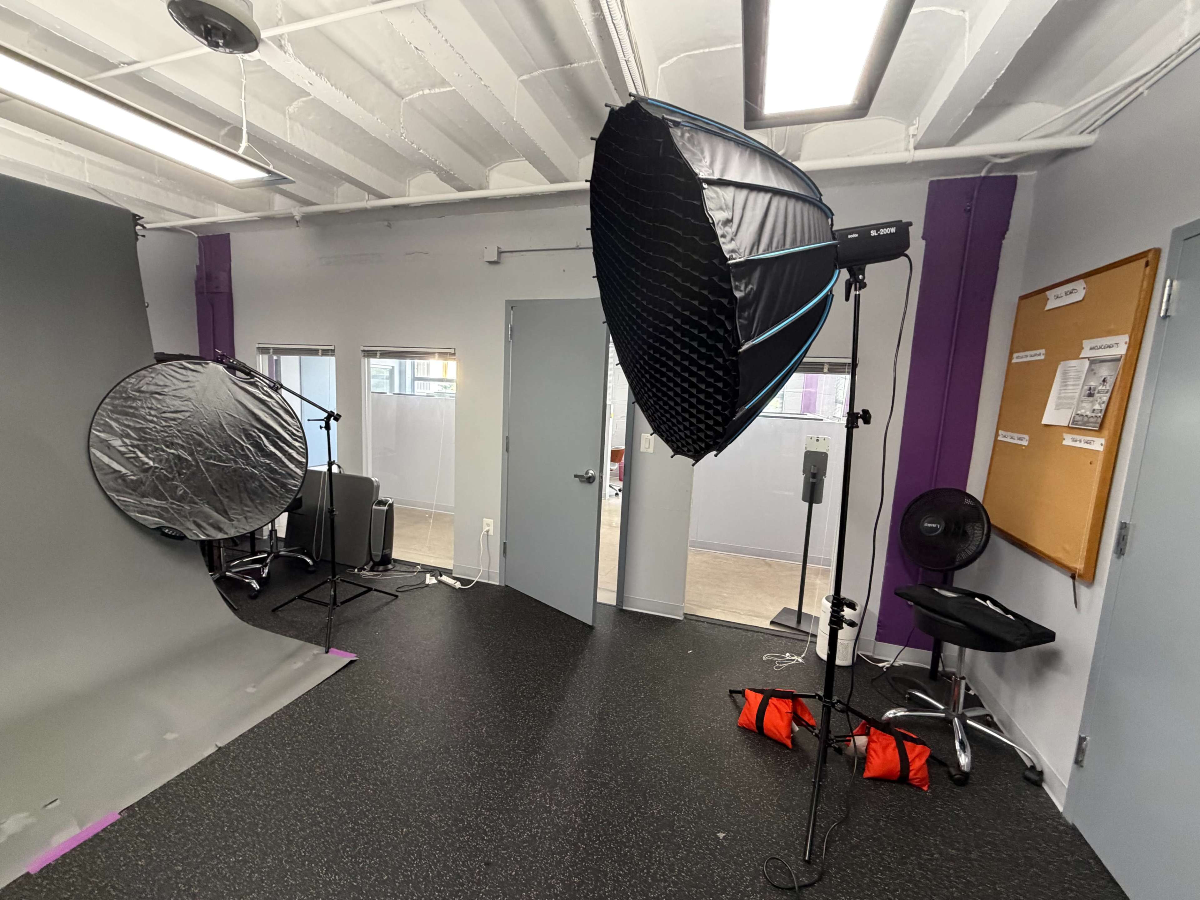 The image shows a photography studio with a gray backdrop, a large softbox lighting setup, a circular reflector, and two desks with chairs.