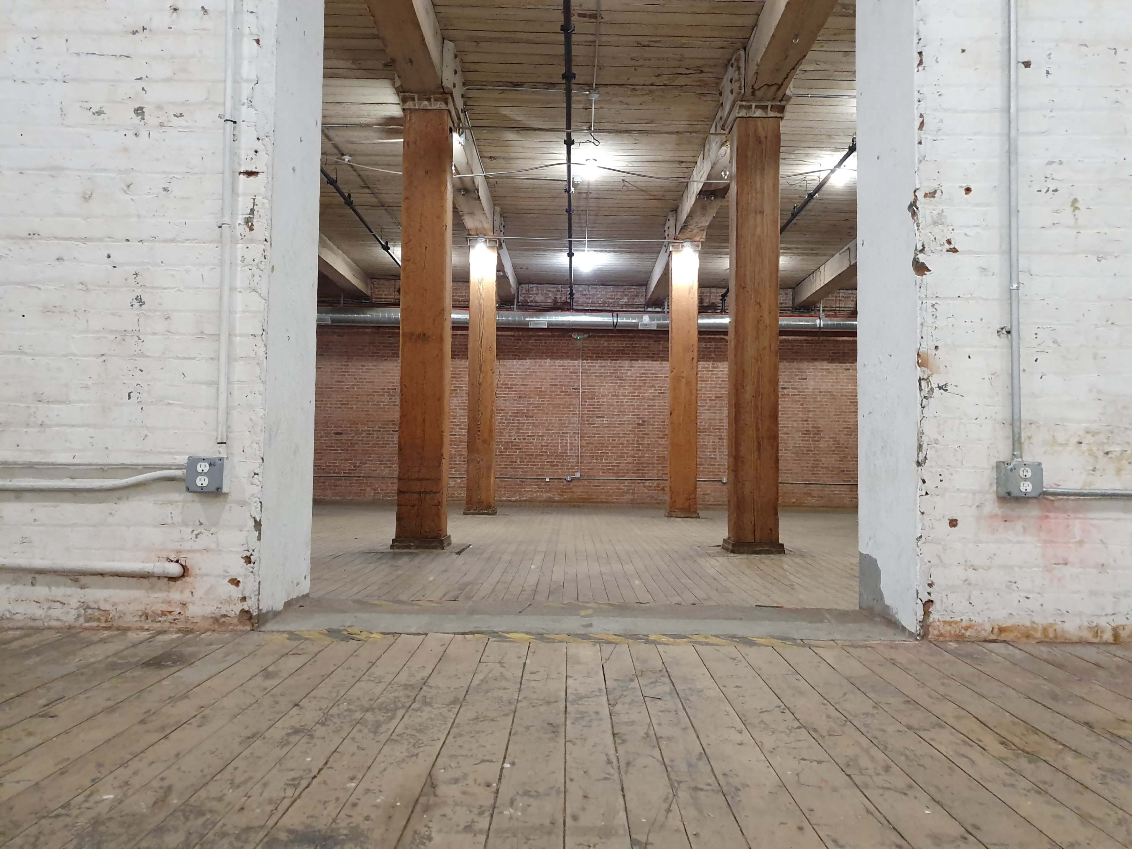 A spacious, empty industrial room with wooden pillars and a wooden floor, surrounded by exposed brick walls.