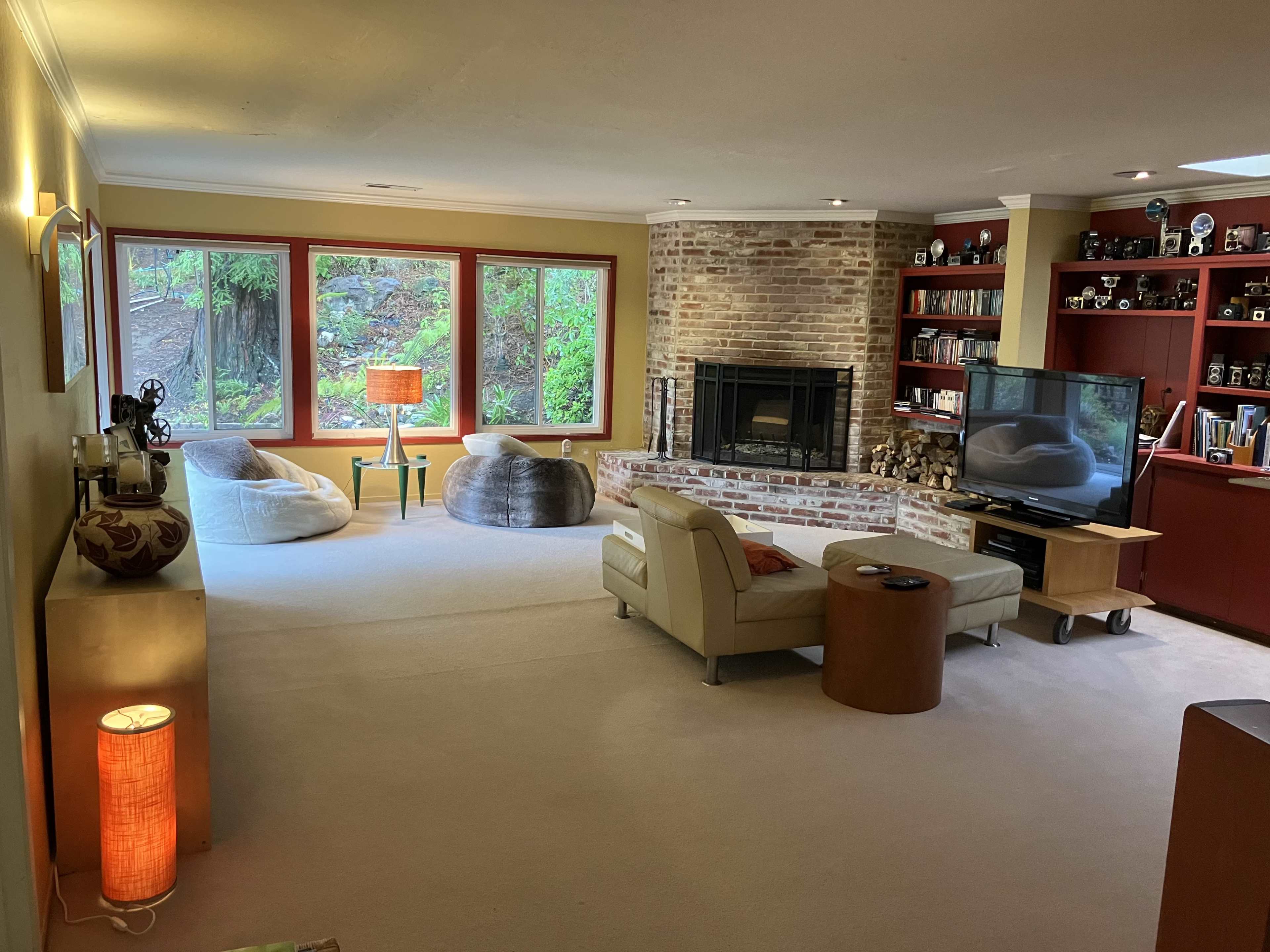 The living room features a pair of bean bag chairs, a brick fireplace, a television on a wheeled cart, and large windows with views of greenery outside.