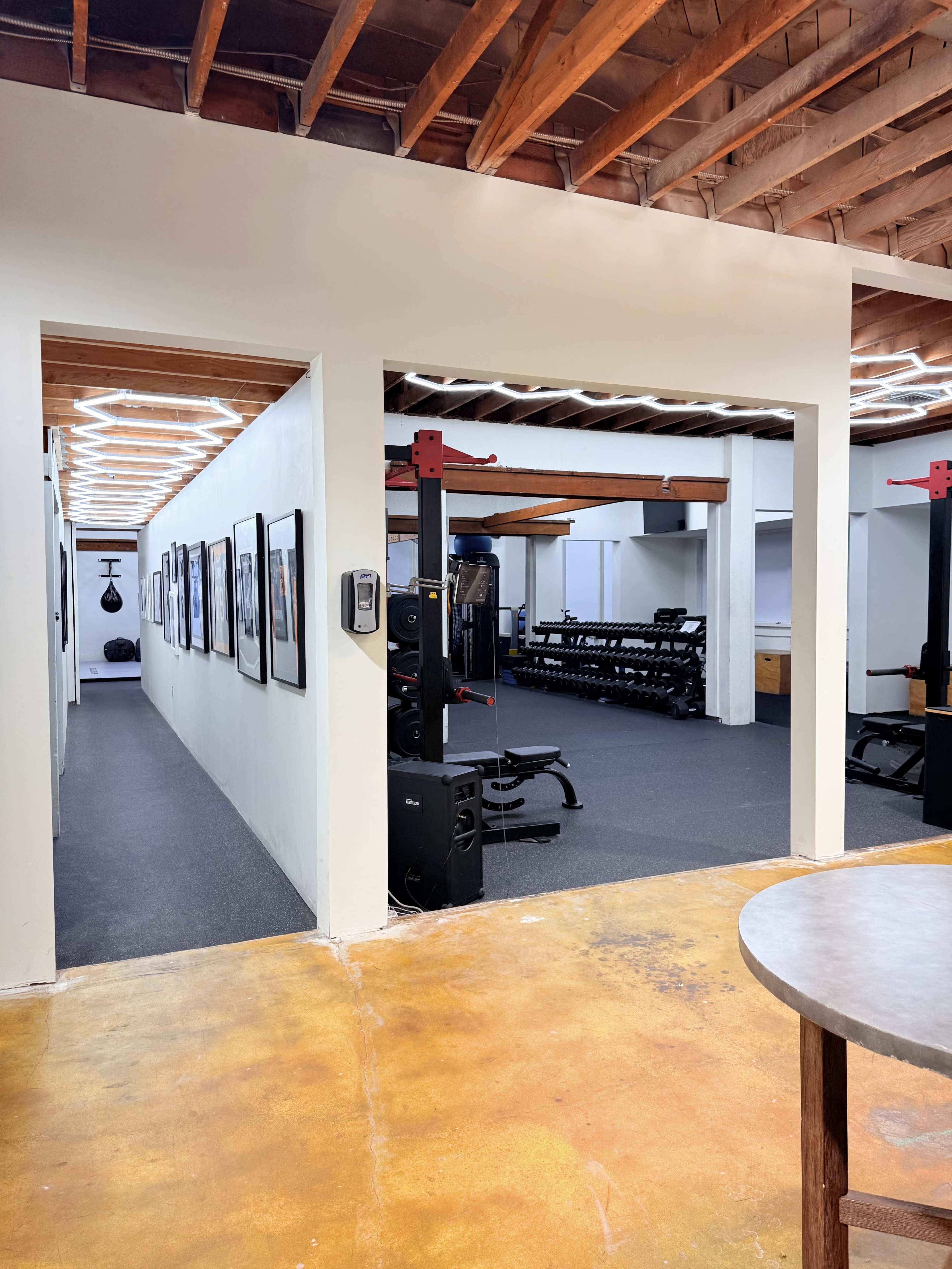 The image shows a modern gym space with workout equipment in the background, framed photographs lining the walls, and a view of a hallway with overhead lighting and a polished floor.