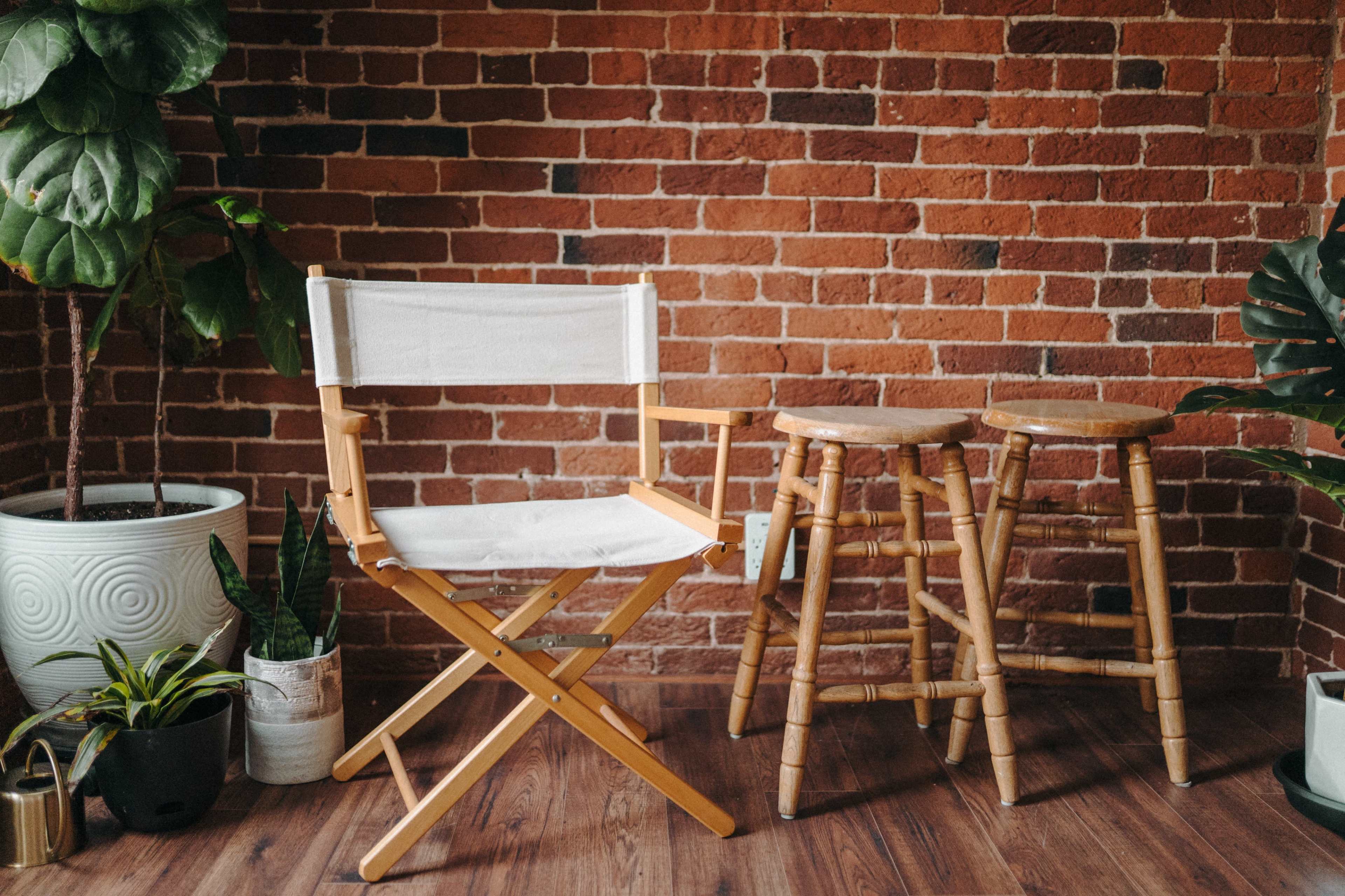 The image shows a wooden director's chair with a white seat positioned beside two wooden stools, set against a brick wall and surrounded by various indoor plants.