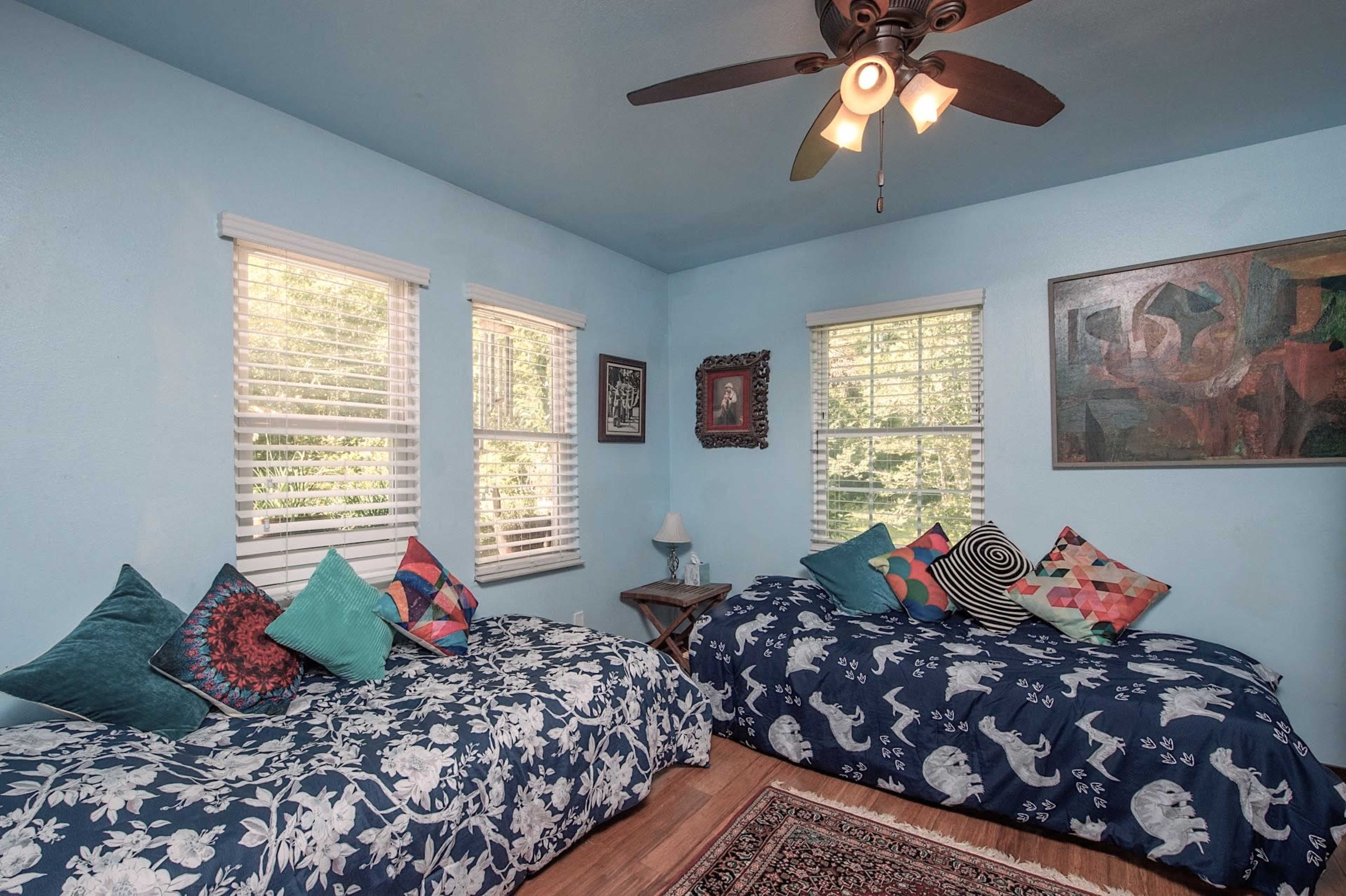 The room features two twin beds with patterned bedding, multiple decorative pillows, and windows that allow natural light to enter.