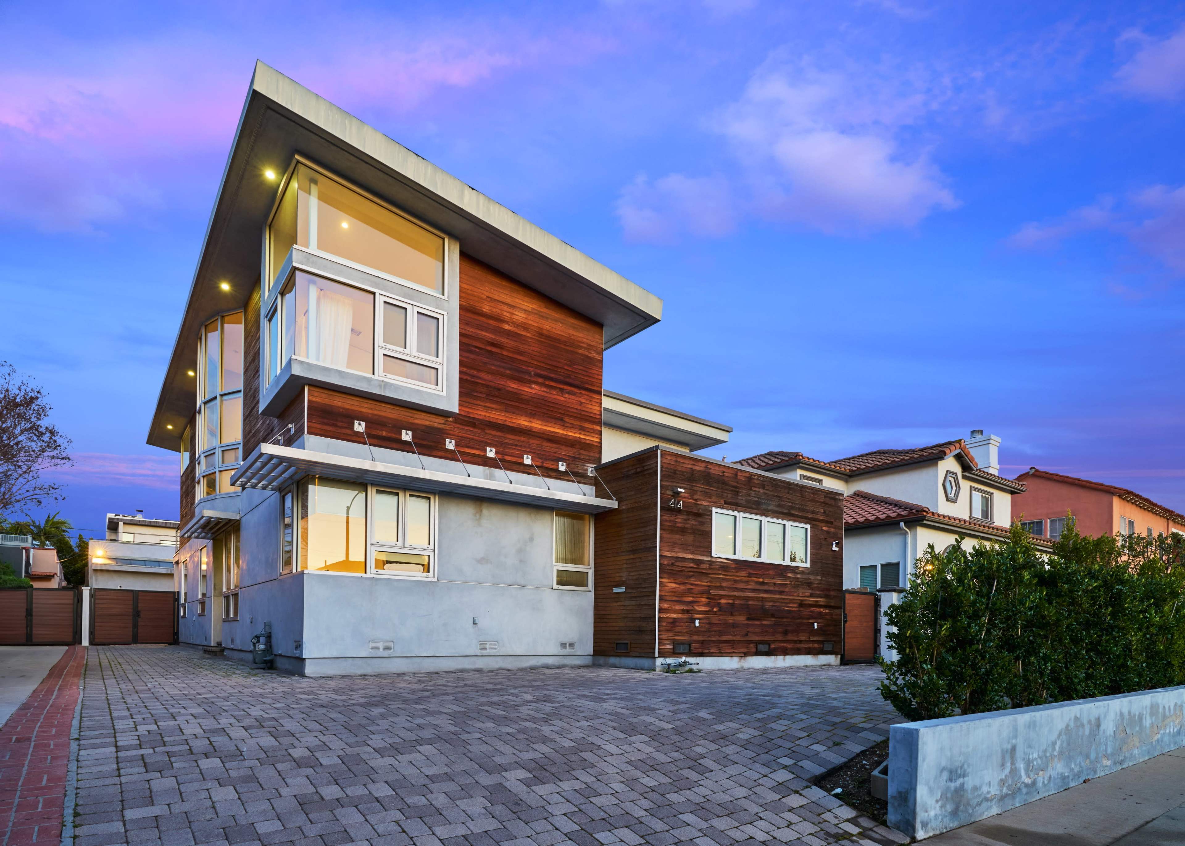 The image shows a modern two-story house with large windows, a wooden facade, and a paved driveway at dusk.