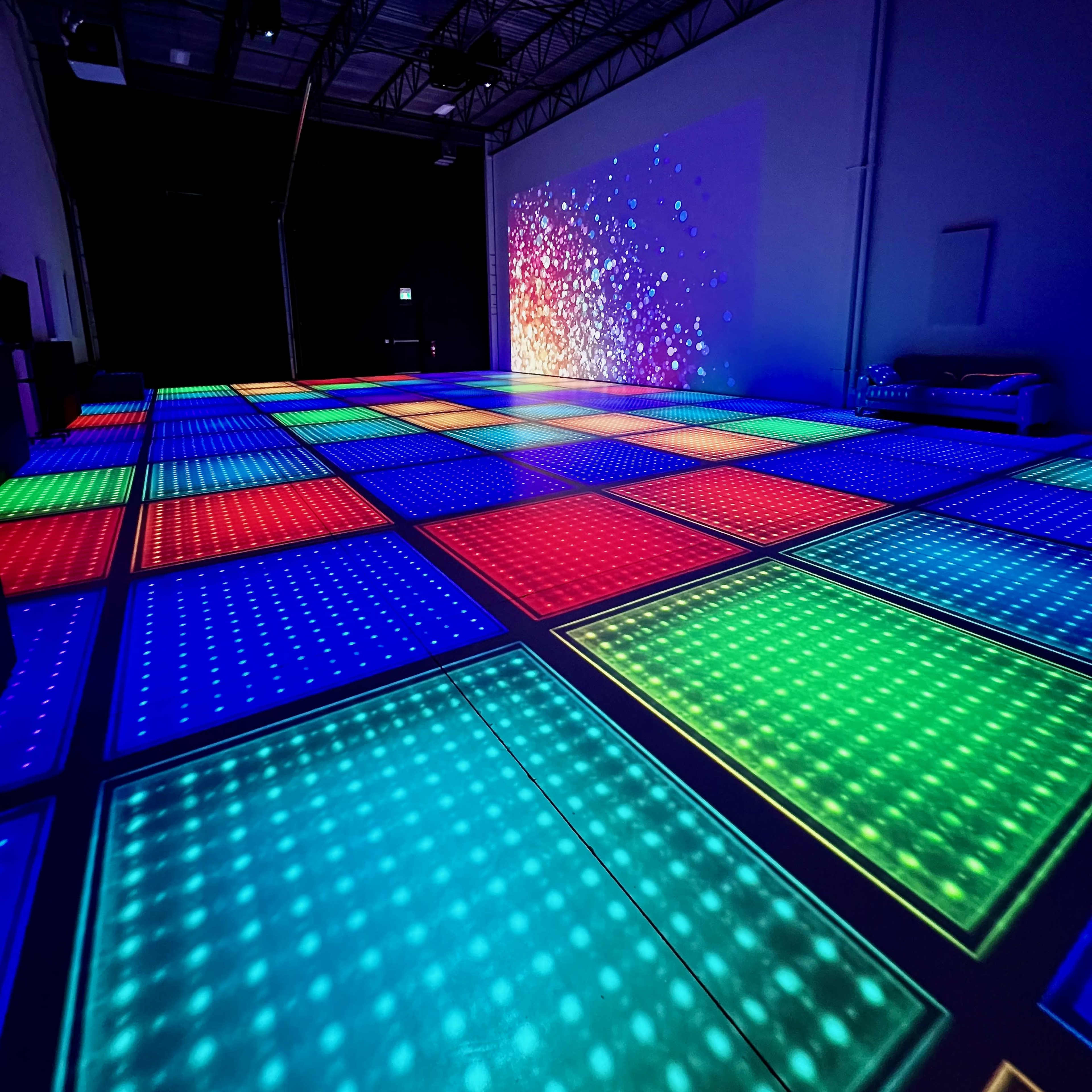 The image shows a brightly lit dance floor composed of multicolored tiles in a spacious indoor area, with colorful projections on one wall.