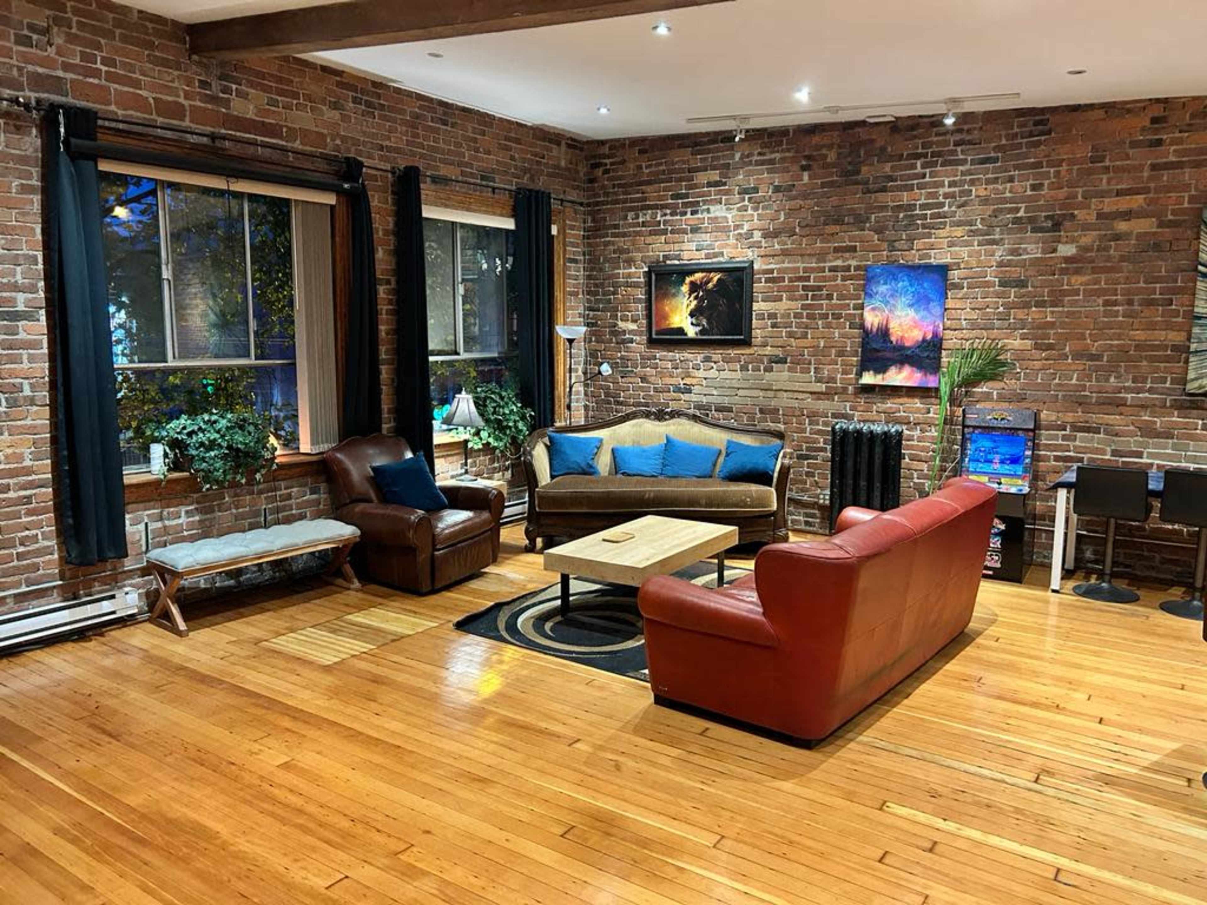 The image shows a living room with exposed brick walls, hardwood floors, and various seating arrangements including a brown leather chair and a red couch.