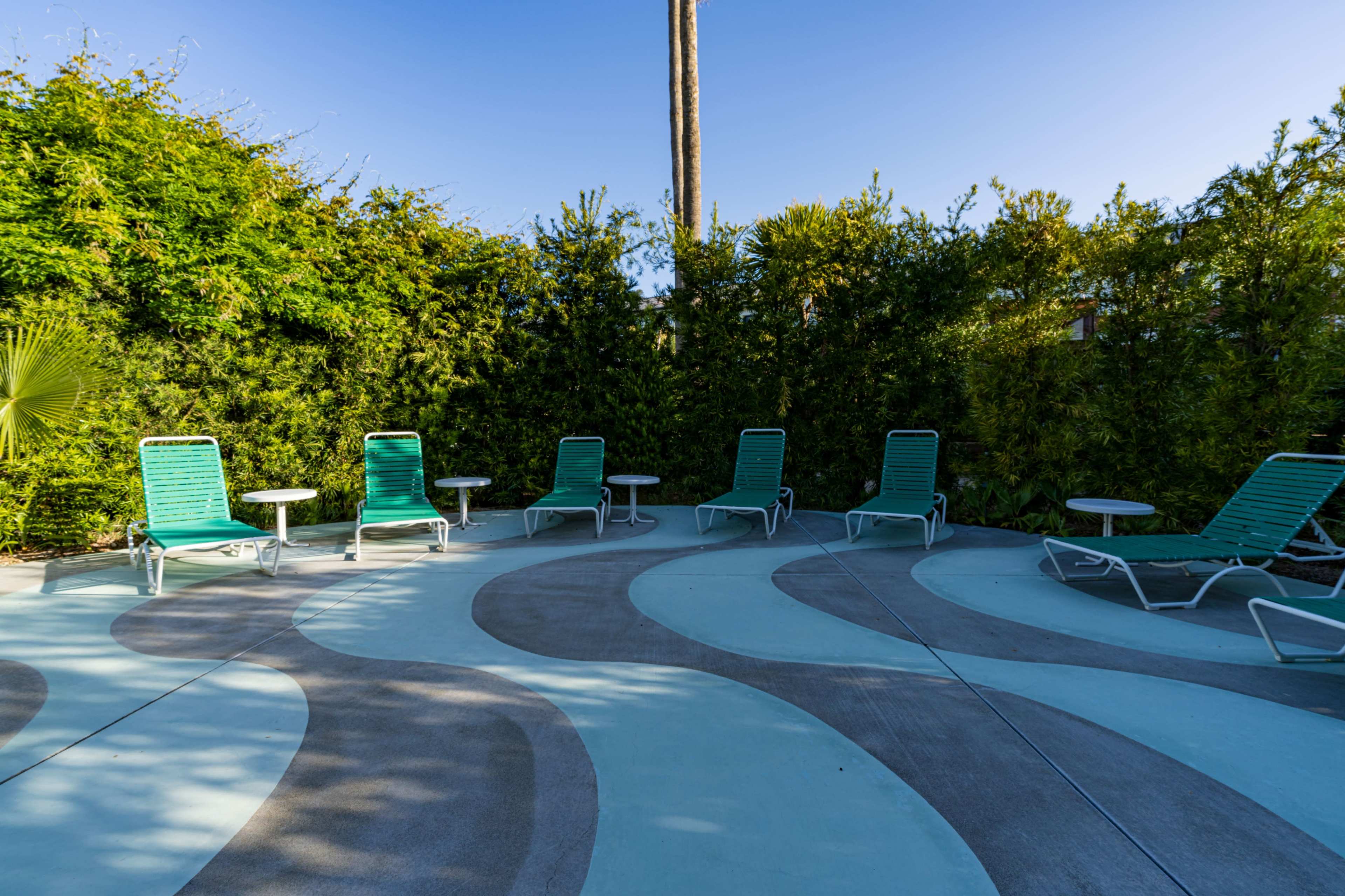 The image shows a patio area with green lounge chairs arranged around small side tables, surrounded by tall greenery.