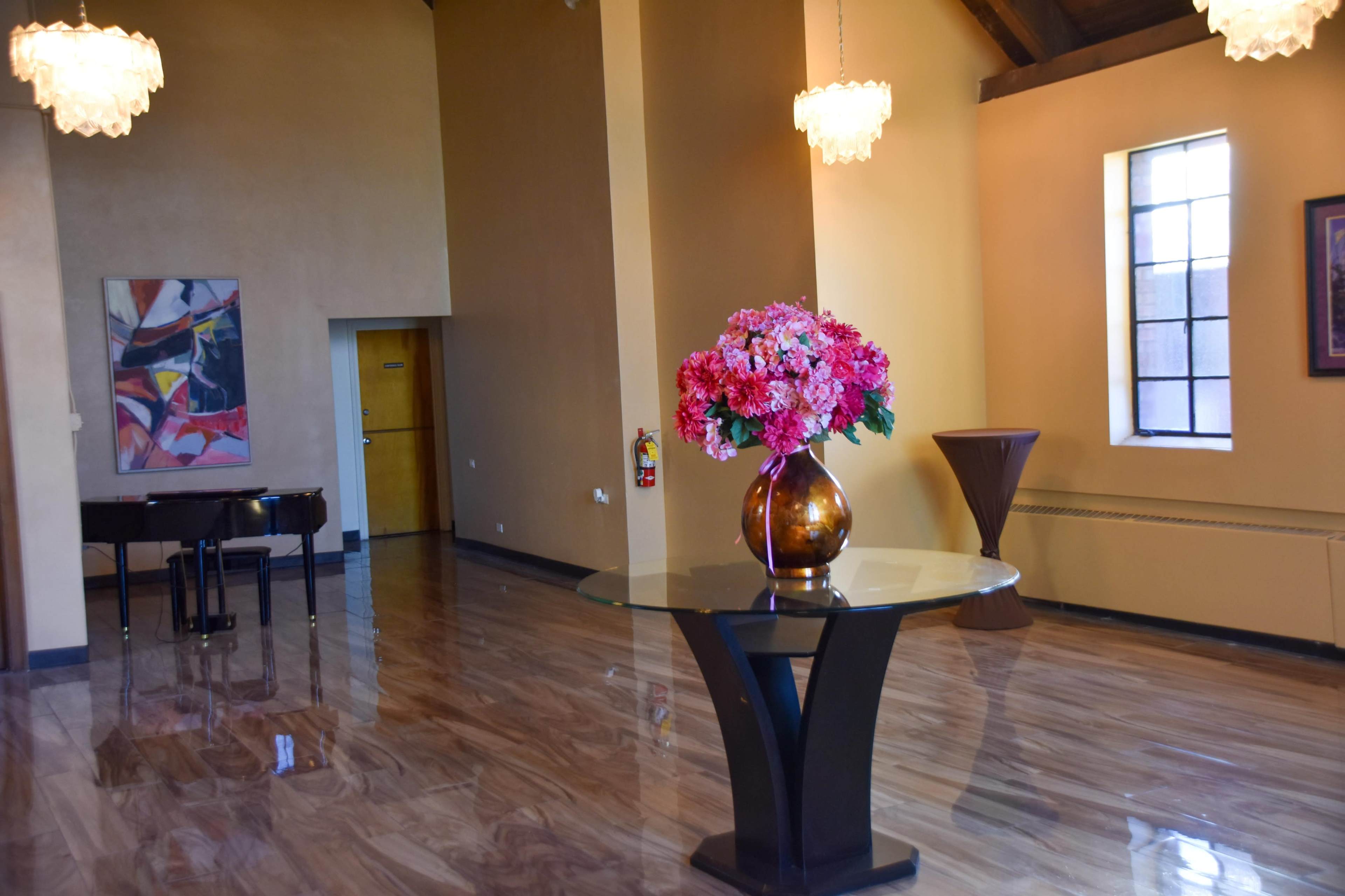 The image shows a spacious interior with polished floors, a large vase of pink flowers on a round table, a grand piano, and two hanging light fixtures.