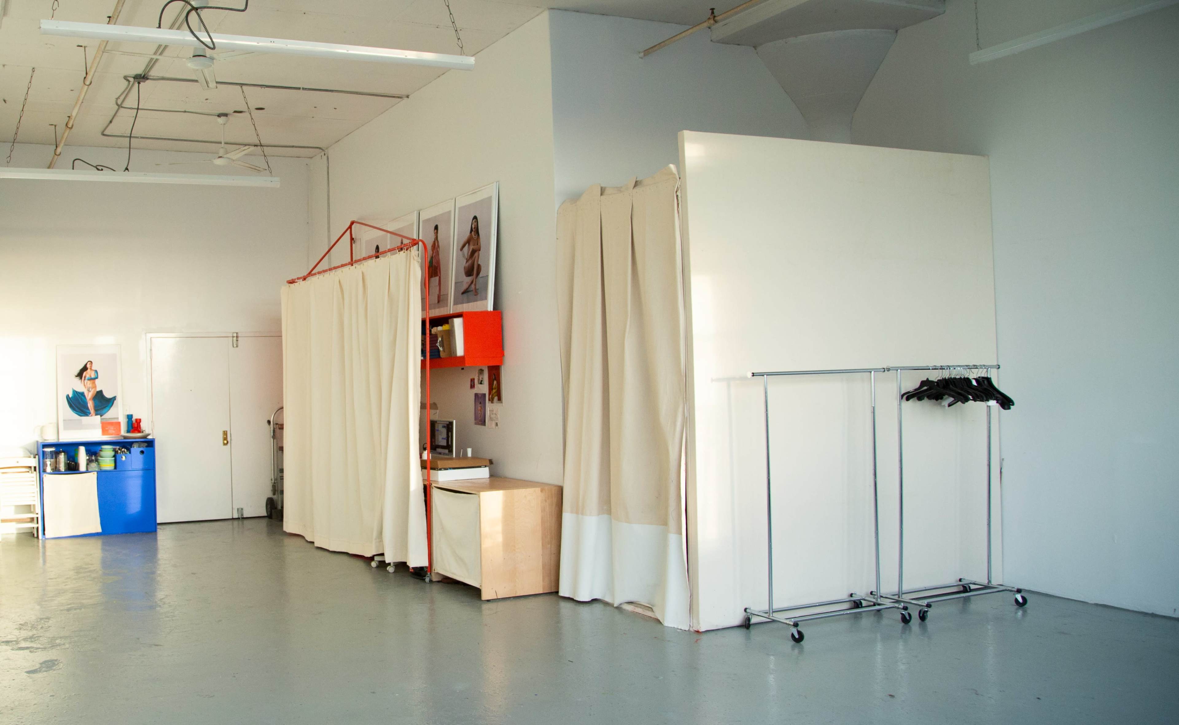 An interior space with light-colored walls, a blue container, a wooden shelf, a clothing rack, and fabric curtains separating different areas.