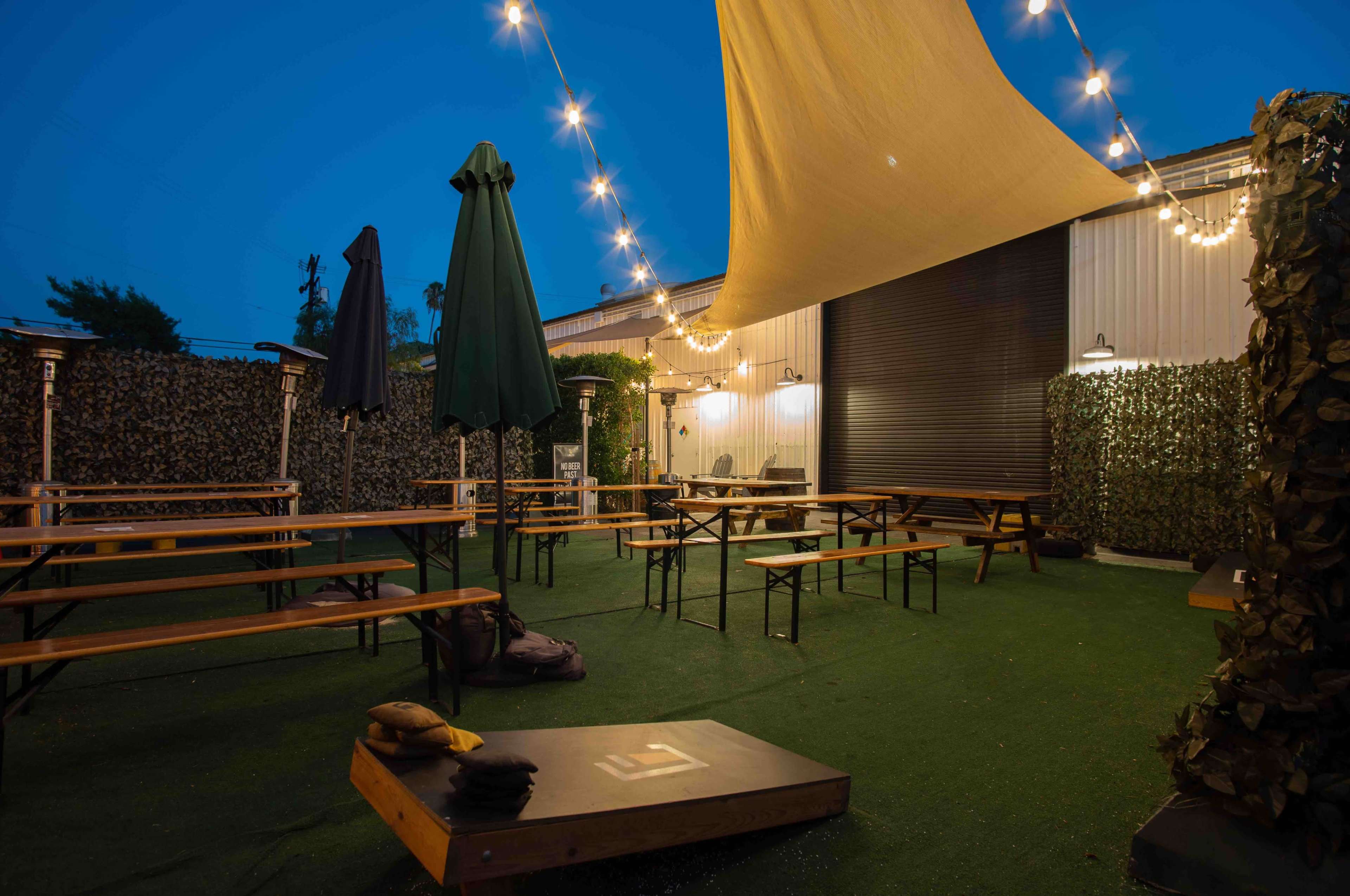The image shows an outdoor seating area with wooden benches and tables, illuminated by string lights and decorated with greenery.