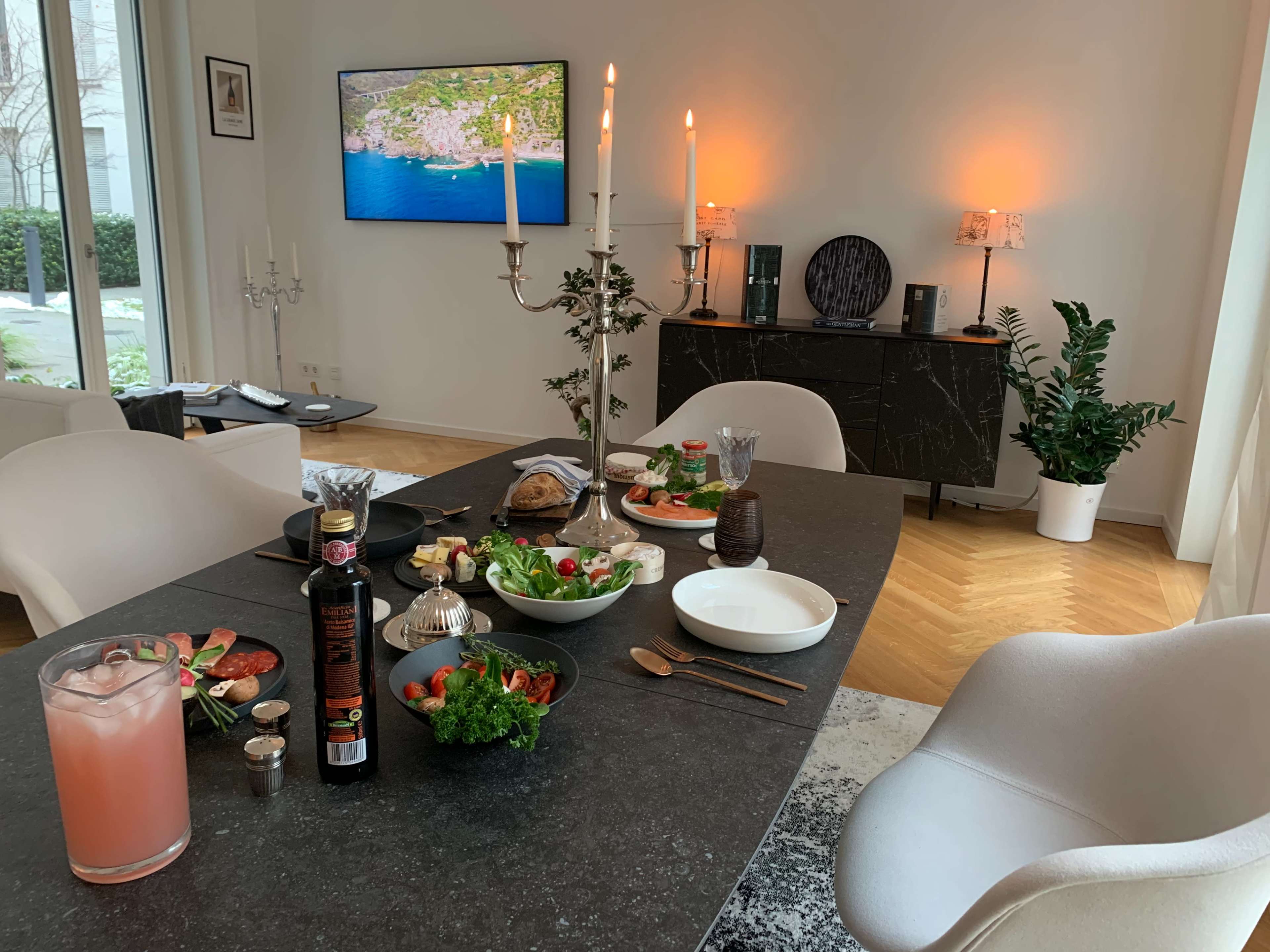 A dining table is set with food and drinks, while a modern living space includes a TV, decorative lighting, and potted plants.