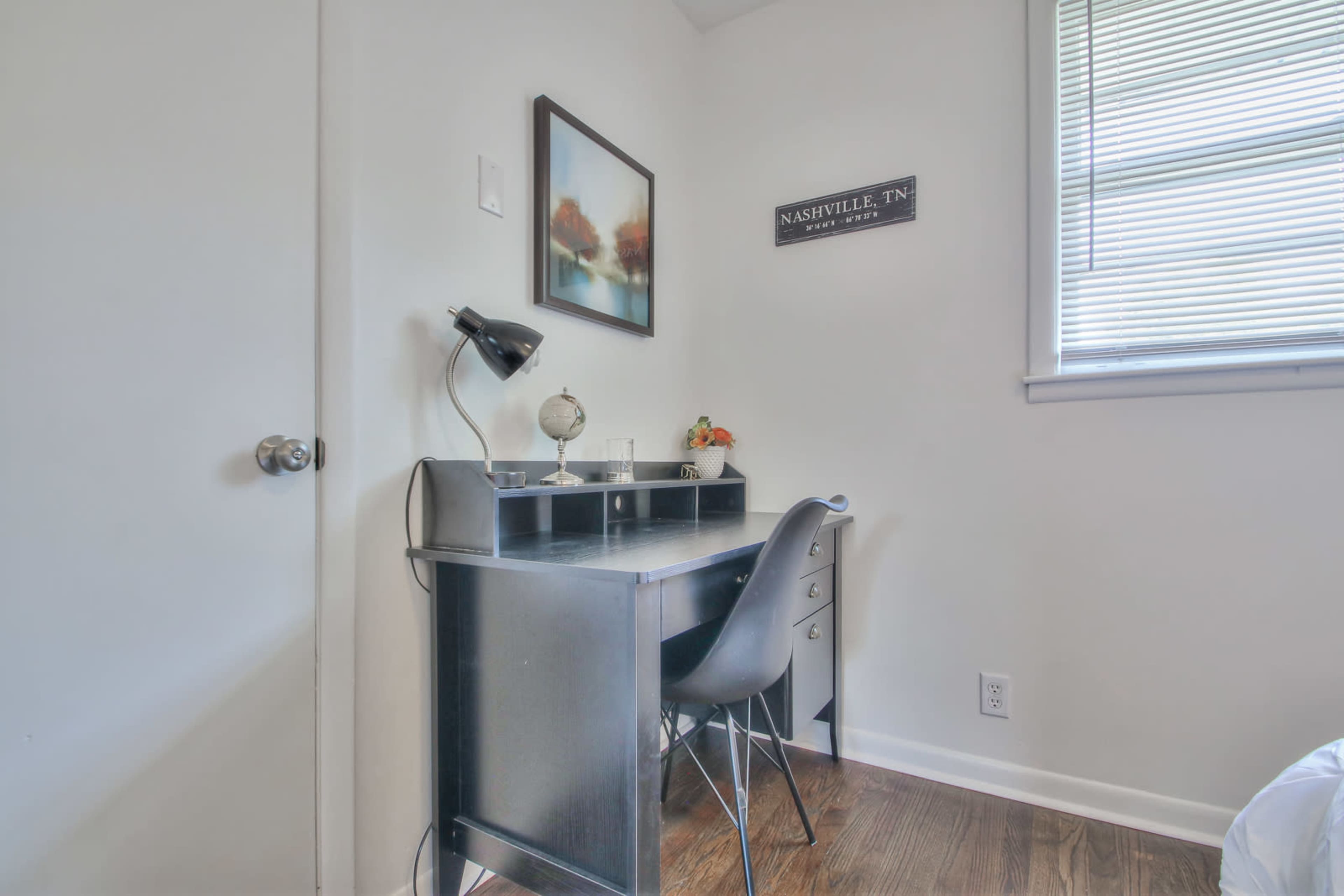 A small desk with a lamp and decorative items is positioned against a light-colored wall in a room with a window.
