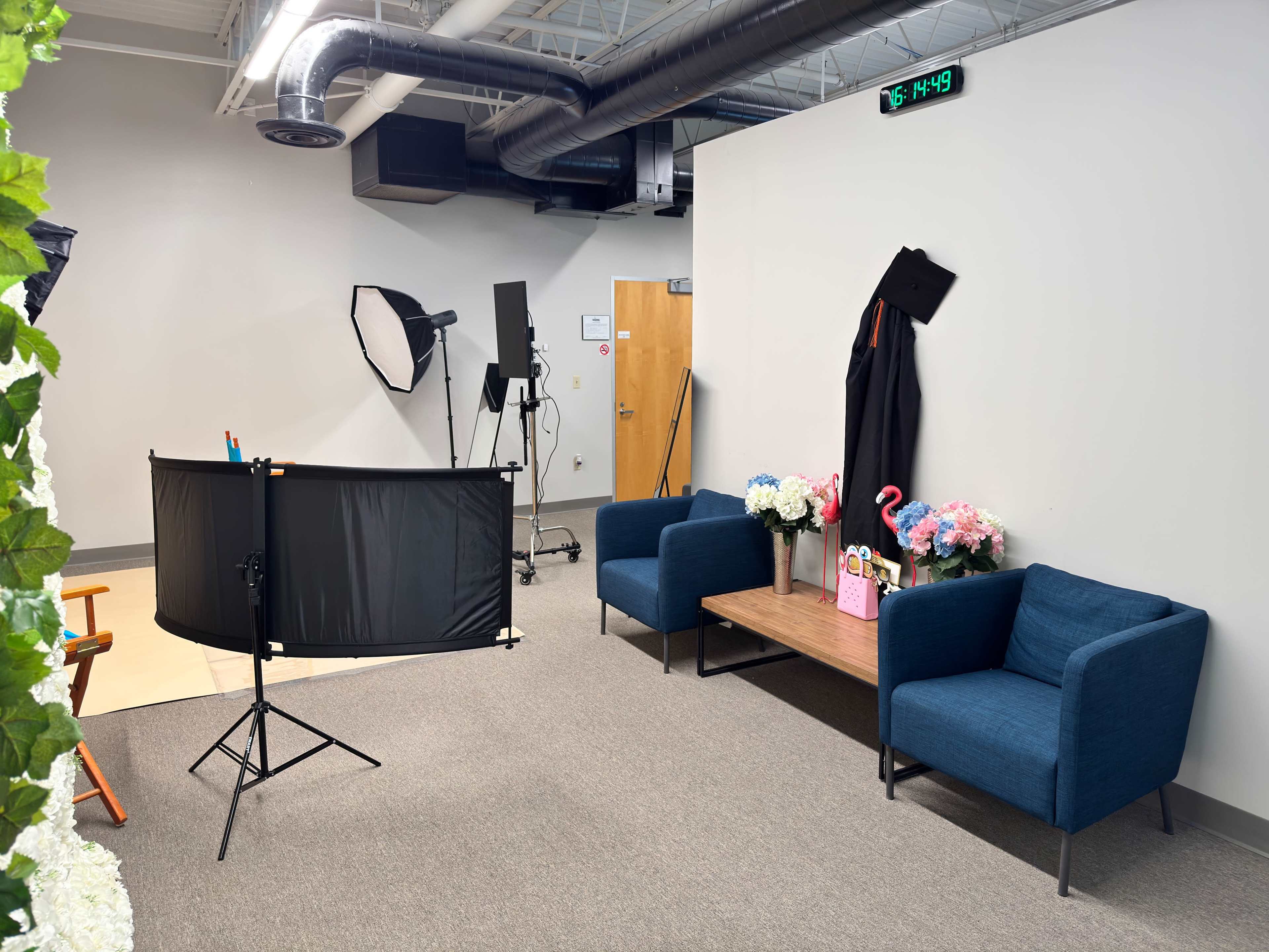 The image shows a studio setup with two blue chairs, a coffee table adorned with flowers and props, photography equipment, and a wall clock displaying 16:49.